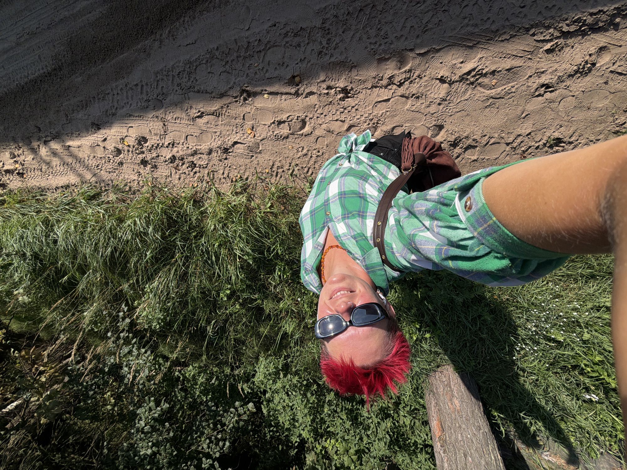 In a high-angle selfie, a person with bright red, spiky hair and sunglasses smiles at the camera. They are wearing a green and white plaid shirt tied at the waist, black pants, and a brown bag slung across their body. They stand on a sandy path with tire tracks and footprints, next to a lush green area with tall grass and foliage. A log lies on the left side of the frame. The scene appears to be an outdoor natural area, possibly a park or trail, in the city of Chaikovsky, Russia, bathed in bright sunlight.