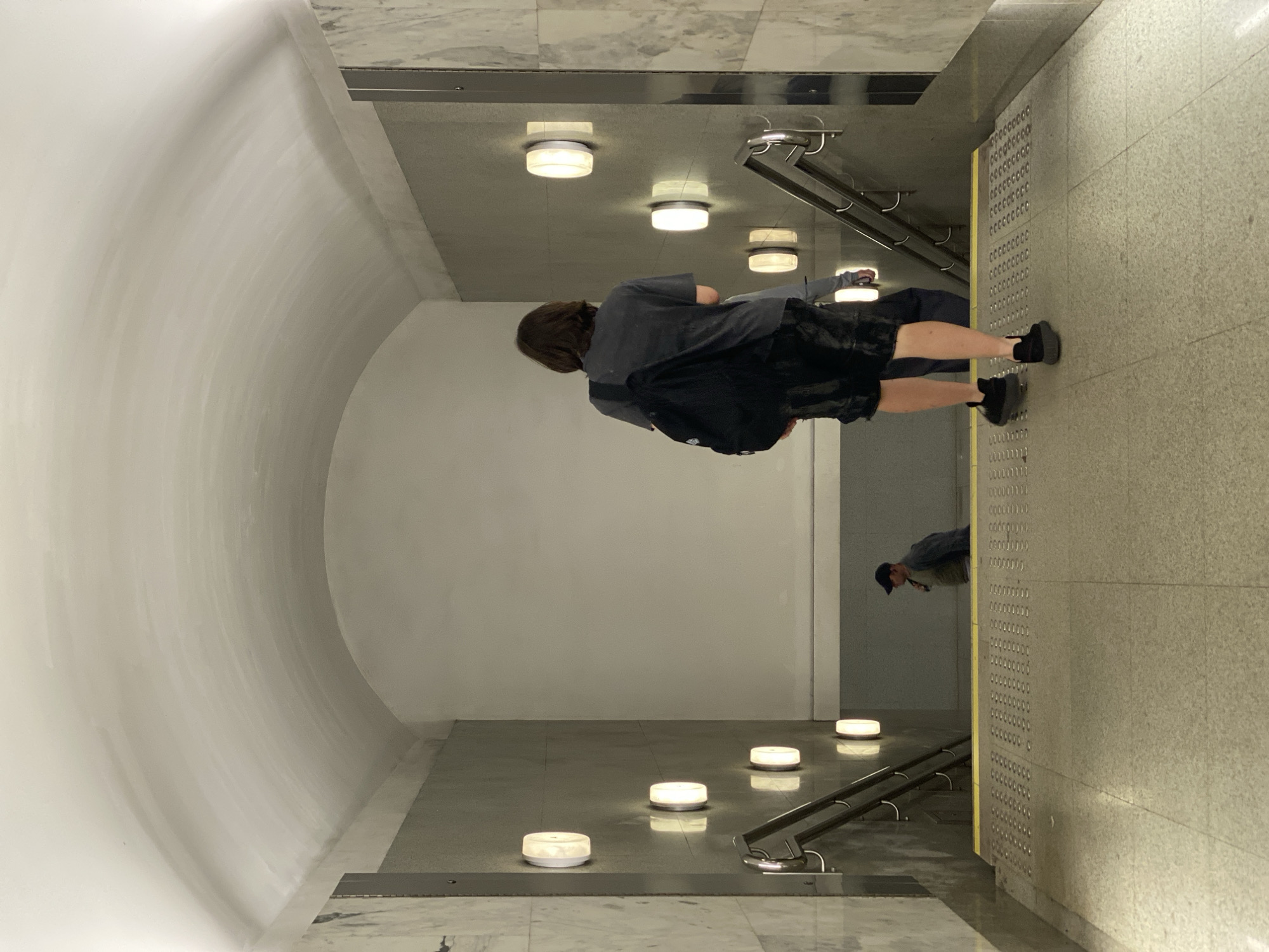 A man with shoulder-length brown hair, wearing a dark grey t-shirt and black shorts, walks up a staircase while carrying a black backpack. He is in the foreground, with his back to the camera. To his left, a man in a dark jacket and a dark baseball cap walks down the stairs, his face obscured. The staircase is part of an underground passage with a vaulted ceiling and marble-clad walls. The walls are lit by oval-shaped wall sconces. The floor is tiled with a pattern of small raised circles, and a yellow tactile warning strip runs along the edge of the stairs. The overall impression is of a subway station or an underpass in Moscow, Russia.