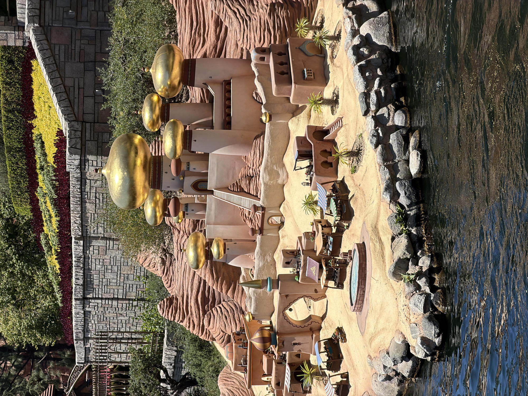 This image captures a meticulously crafted miniature scene resembling a desert oasis village, likely a themed attraction. The architectural style is reminiscent of Middle Eastern or Arabian designs, with sand-colored buildings, arched doorways, and numerous golden domes topping spires and structures. The foreground features a body of dark, rippling water, with rocky shorelines. Behind the village, sculpted red rock formations rise, creating a layered backdrop. Lush green vegetation and a vibrant, multi-colored floral display (red, yellow, and green) adorn the top of a stone wall in the upper portion of the image. The scene is bathed in bright, natural sunlight, indicating it's daytime. This setting is characteristic of Disneyland Park in Anaheim, California, specifically the "Aladdin's Oasis" or a similar Fantasyland attraction. There are no people or animals visibly depicted in the scene.