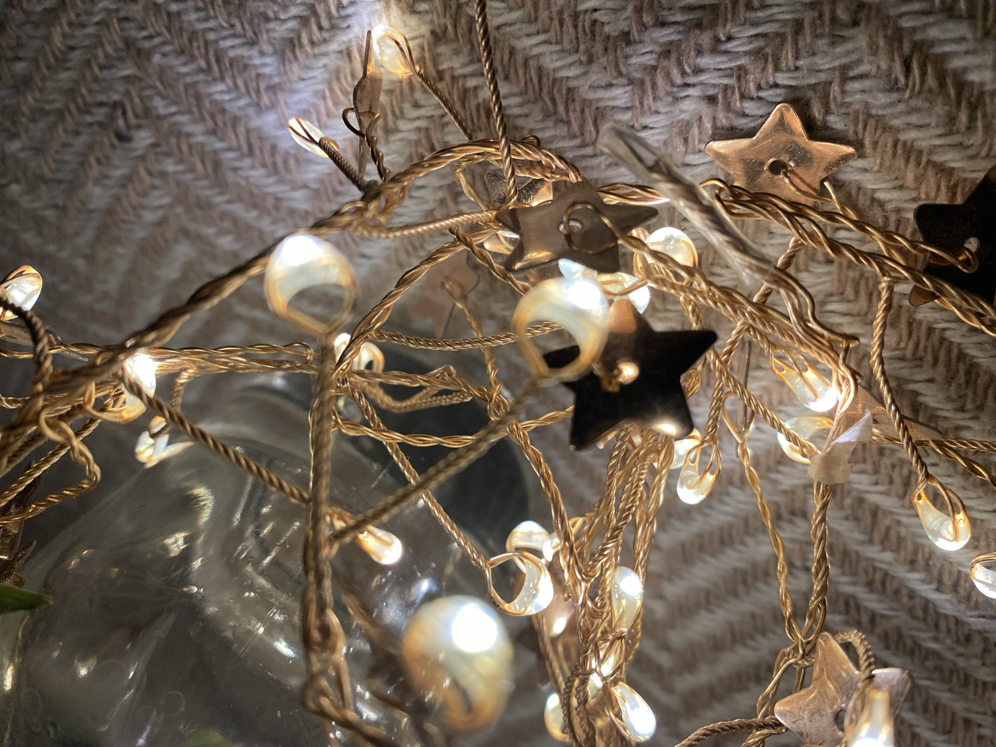 The image depicts a close-up view of decorative lights. The lights consist of small, teardrop-shaped bulbs and star-shaped ornaments. The lights are intertwined with a thin, gold-colored wire. The background appears to be a textured, woven fabric or material. A partially visible, reflective glass object is present on the left side of the image. There is no evidence to suggest the location is Chaikovsky, Russia. There are no people visible in the image.