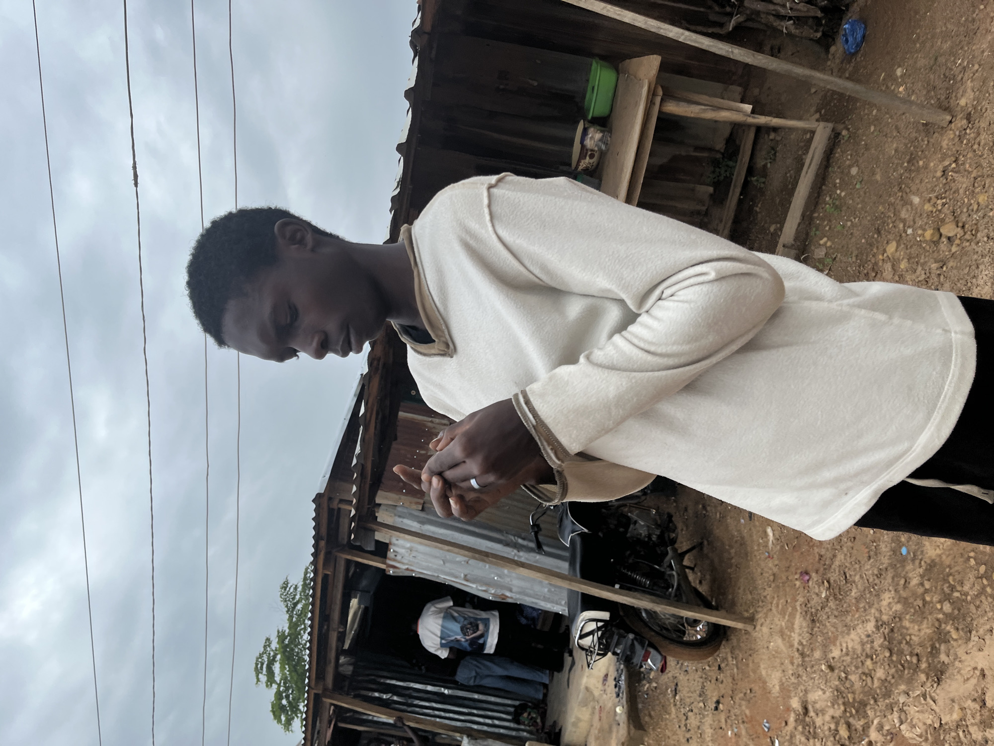 A young Black male stands in the foreground of the image, looking down at his hands which are clasped together. He is wearing a light-colored long-sleeved shirt and black pants. His hands are positioned such that a ring is visible on his left ring finger. In the background, a simple open-front structure with a corrugated metal roof and walls made of wood and corrugated metal is visible. A motorcycle is parked to the left of the structure, and a wooden table with two containers sits to the right. The ground is dirt and sparsely covered with small stones and debris. The sky is overcast and cloudy. The scene depicts a street in a densely populated area, likely a market or residential neighborhood, which aligns with the typical streetscape of Jos, Nigeria, characterized by informal structures and unpaved roads.