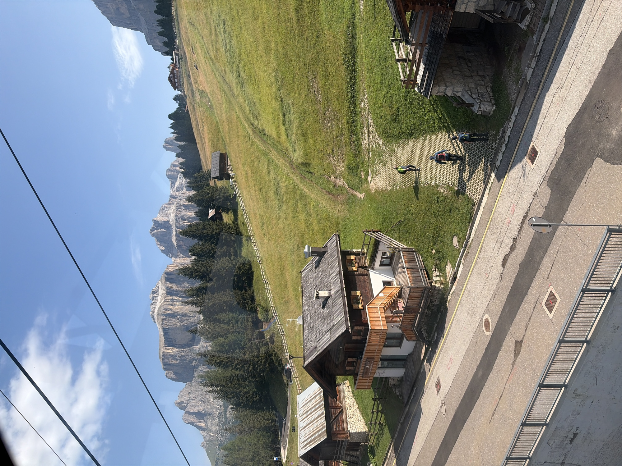 In Canazei, Italy, a scenic mountain landscape unfolds under a clear blue sky with scattered clouds. Cable car lines stretch across the frame, hinting at the mode of transport for visitors. The foreground features a paved road with a grey metal railing, transitioning to a grassy slope dotted with wooden chalets. Several people are observed walking on a paved path winding up the hillside, some carrying backpacks. The chalets, built in a rustic alpine style with wooden facades and shingled roofs, are nestled amongst evergreen trees. In the background, imposing rocky mountain peaks dominate the horizon, their rugged surfaces illuminated by the sun. The scene captures a serene mountain village setting during daylight hours.