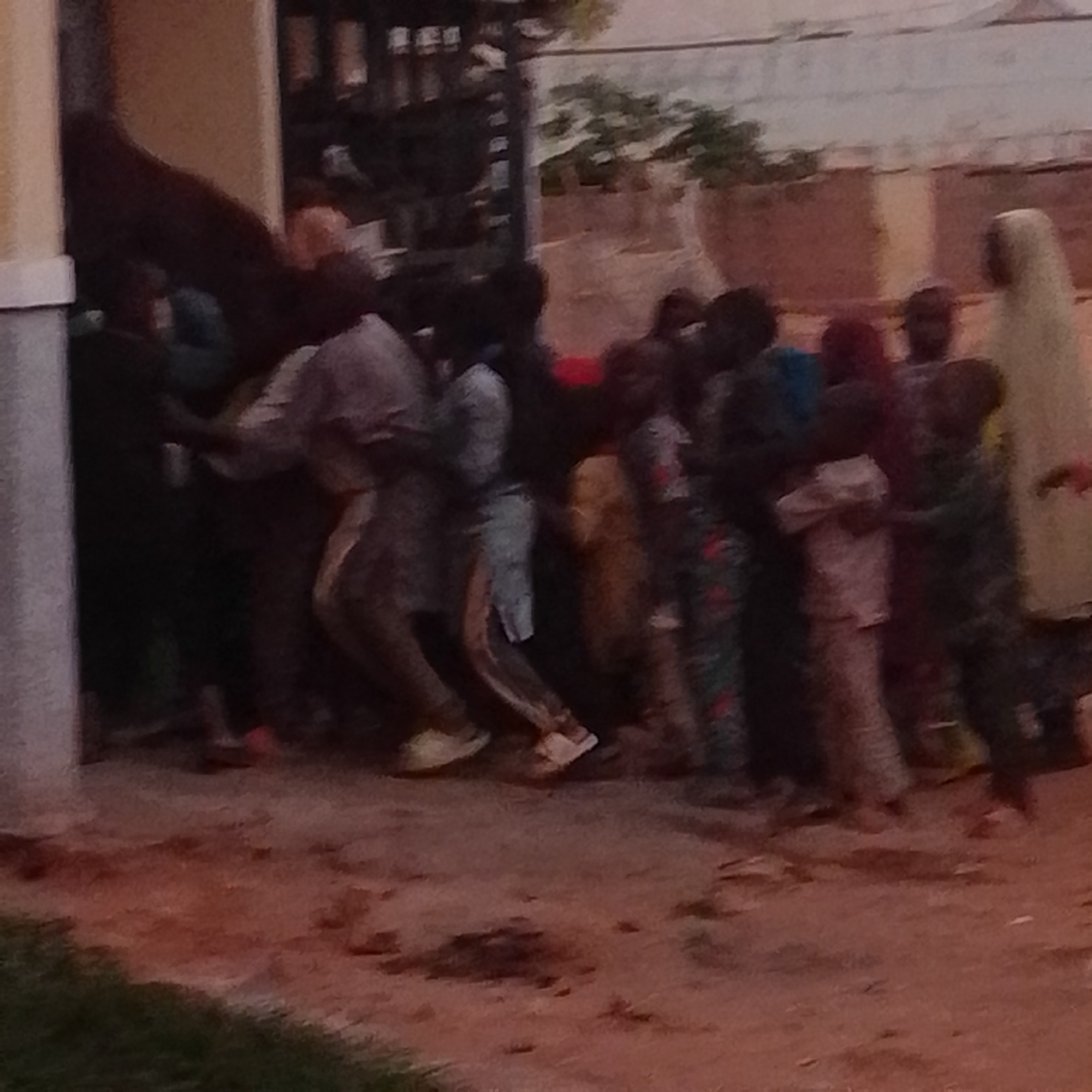 This media file depicts a group of people in what appears to be an outdoor setting in Gombe, Nigeria. The subjects are a mixture of individuals, predominantly dressed in traditional African attire, with some wearing headscarves and flowing garments. The scene is dimly lit, suggesting it might be late afternoon or early evening. The ground is a reddish-brown dirt, with some patches of green grass visible in the foreground.
The people are clustered together, appearing to be engaged in some form of gathering or activity. Some individuals are facing away from the camera, while others are partially visible. The overall impression is that of a community event or a social interaction. The background shows a building with a textured, possibly brick, facade and a light-colored roof or wall. There are also some trees and vegetation visible in the distance, hinting at a semi-urban or rural environment.
The image is slightly blurry and low resolution, which makes it difficult to discern specific facial expressions or finer details. However, the gathering of people suggests a sense of community and shared experience. The lighting and the attire of the individuals point towards a location in Nigeria. No visible text is present in the media file.