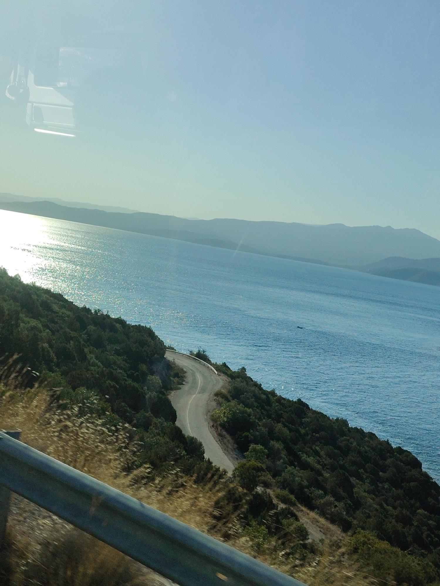 The image depicts a scenic coastal road winding through a green, hilly landscape overlooking a vast expanse of the Aegean Sea. The sun glints off the water, creating a shimmering effect. In the foreground, a metal guardrail runs along the edge of the road, suggesting a viewpoint from a moving vehicle. The road itself is asphalt, with a white line marking its edge. The vegetation is dense and appears to be a mix of trees and shrubs. In the distance, hazy mountains form a layered backdrop against the clear blue sky. The sky transitions from a pale blue near the horizon to a deeper blue overhead. The image is likely captured from a car driving along the coastal highway near the Municipal Unit of Pelasgia, Greece, known for its stunning sea views and winding roads. There are no people or animals visible in the frame.