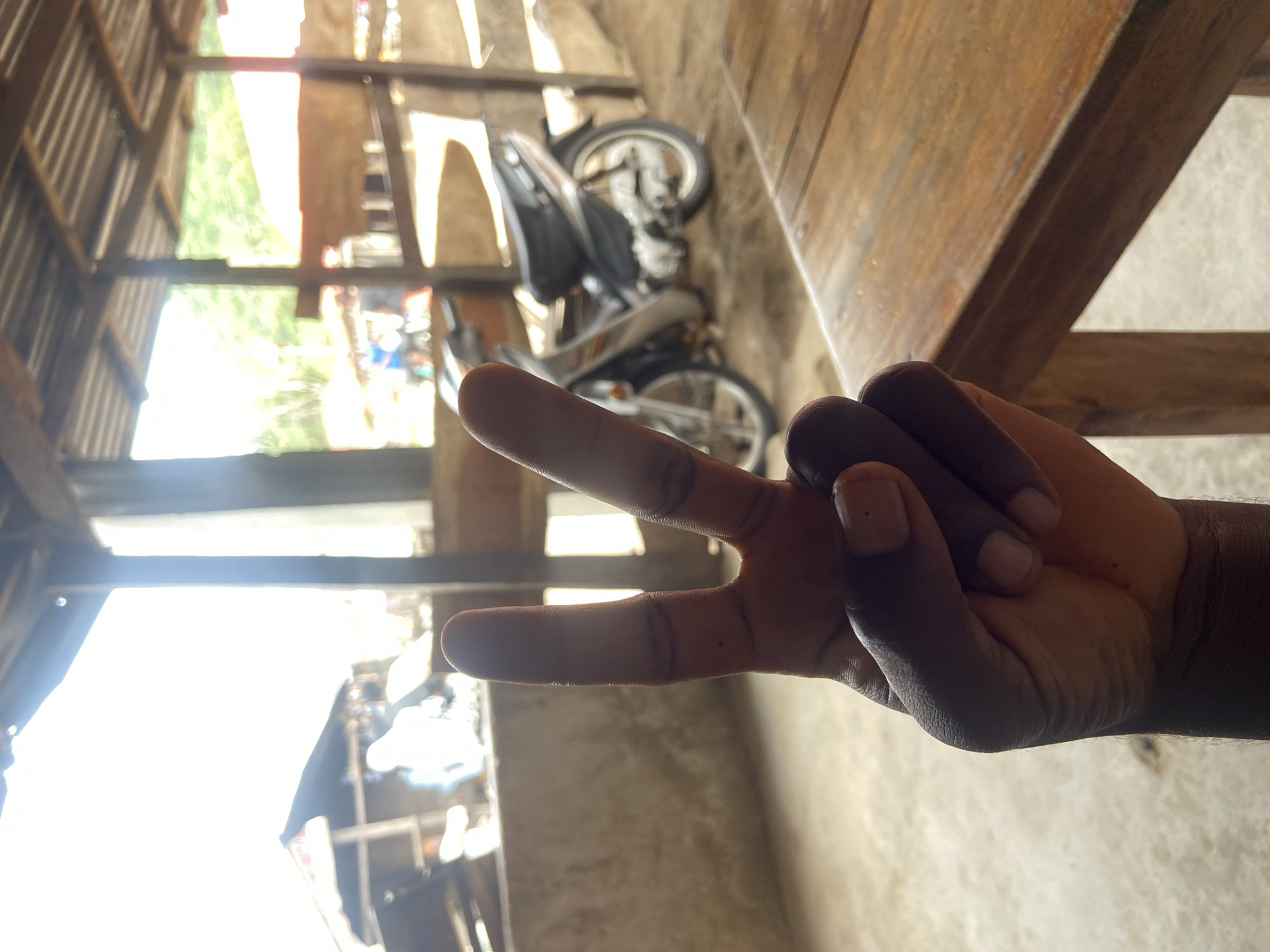 The image depicts a hand making a peace sign against a blurred background. In the background, a motorcycle is partially visible, parked under a roofed structure. Outside the structure, glimpses of buildings and sunlight suggest an outdoor environment. The image was taken in Mararaba, Nigeria.