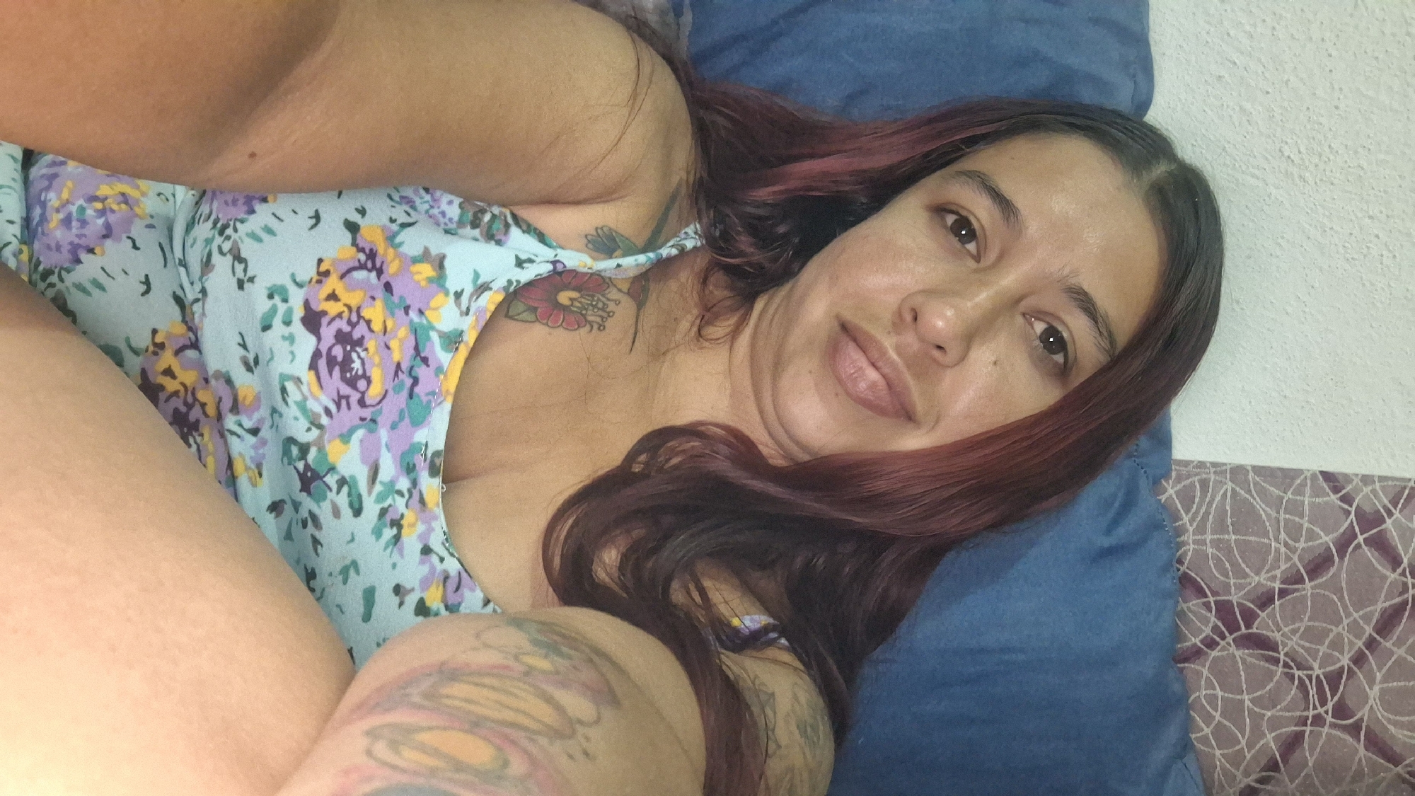 A woman is lying on a blue pillow and a purple and white patterned surface. She has brown hair with red highlights, light brown skin, and a tattoo on her shoulder and arm. She is wearing a light blue dress with purple and yellow floral patterns. The background consists of a white wall and part of the patterned surface. There are no indications of the location being Vallecito, Venezuela.