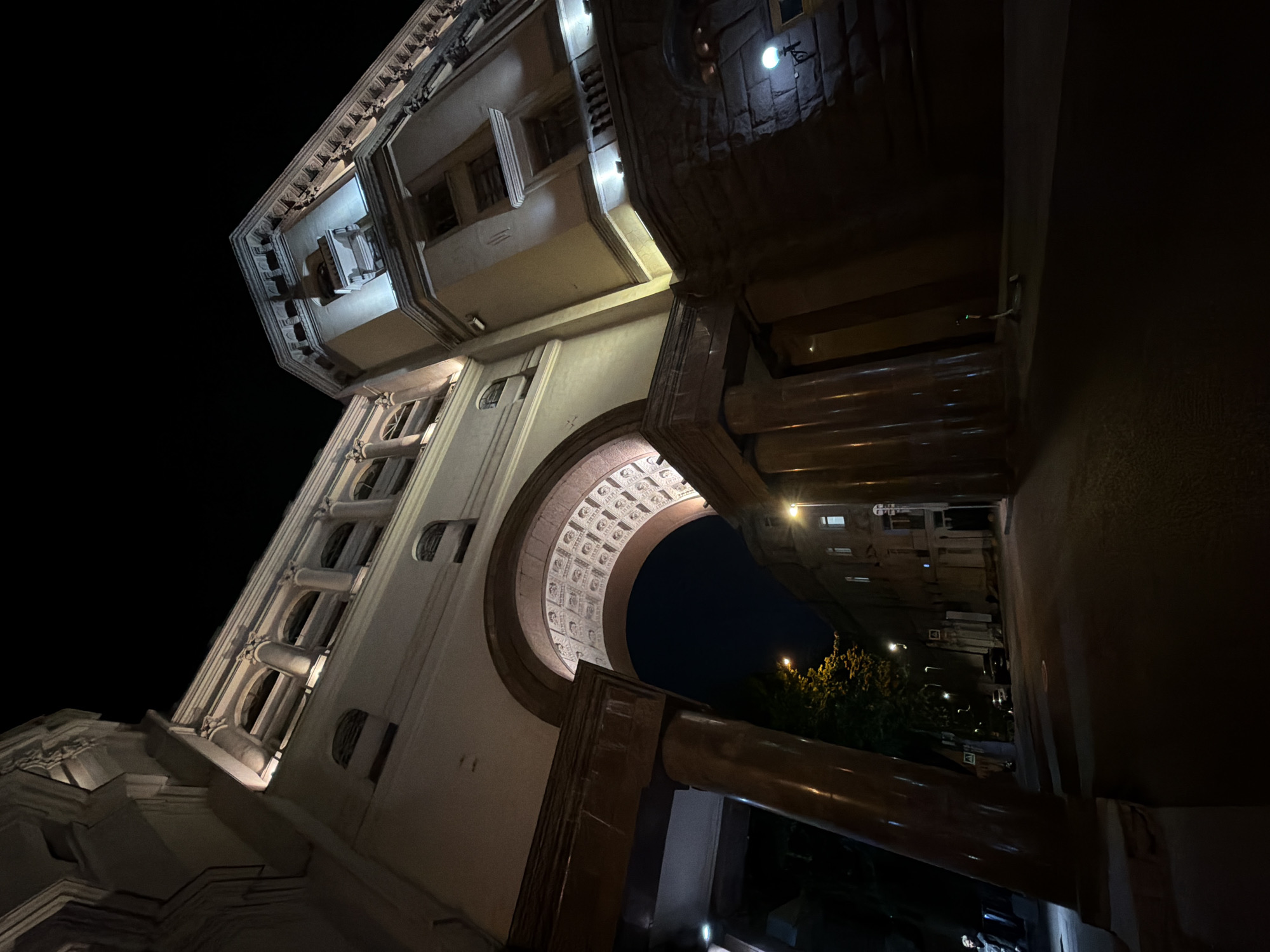 The image captures an architectural perspective from below, focusing on an archway in Moscow, Russia. The archway is part of a building illuminated by artificial lighting, showcasing its intricate design. Beyond the arch, the night sky is visible, along with distant buildings and streetlights. The perspective is angled, emphasizing the height and grandeur of the architecture.