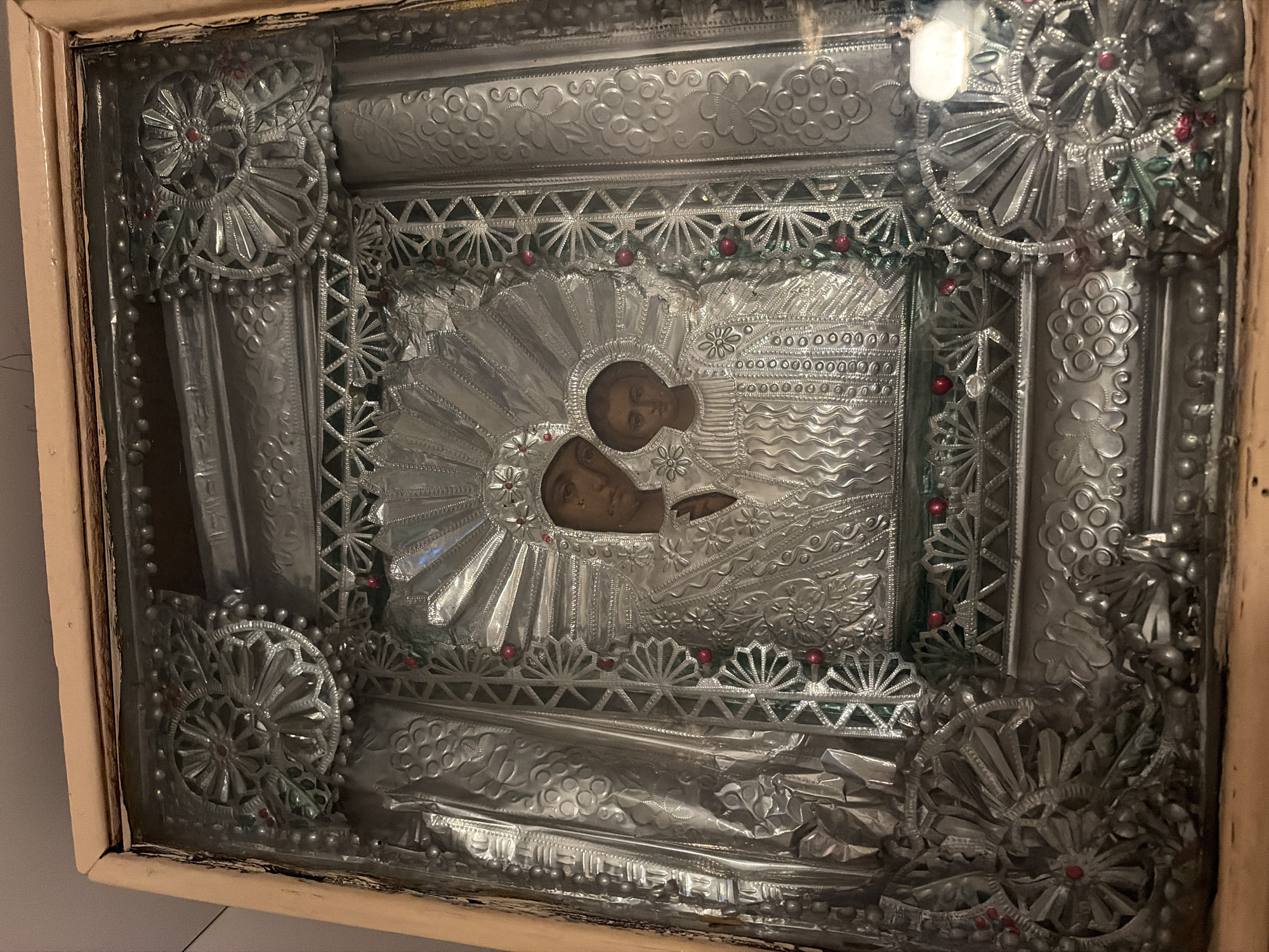 This is a close-up, eye-level shot of an ornate, silver-toned icon of the Virgin Mary and child Jesus, housed within a wooden frame and protected by glass. The icon features intricate embossed detailing, with the figures of Mary and Jesus depicted centrally, adorned with jeweled halos and ornate clothing. The surrounding metalwork is richly decorated with geometric patterns, floral motifs, and small red embellishments, resembling small beads or gems. The overall impression is one of religious significance and craftsmanship. There is no discernible indication of the city of Tambov, Russia, within the image itself.
