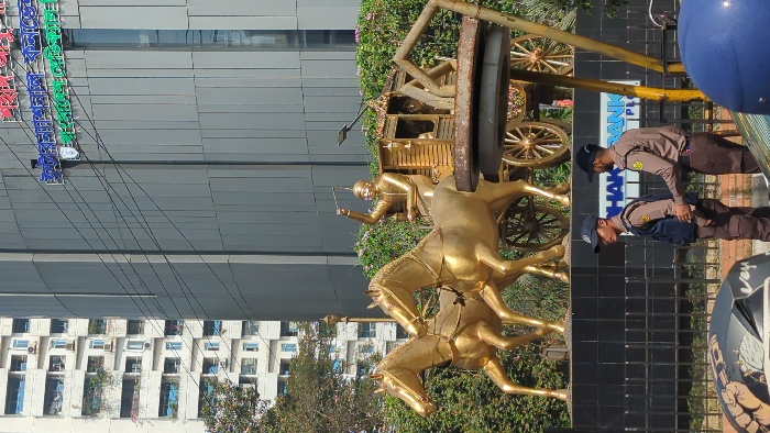 A golden statue depicts a chariot pulled by two horses with a driver holding reins. Behind it, is a wooden cart with spoked wheels. Two men in uniform stand in front of a dark building. A building with a colorful illuminated sign is visible in the background. Another building with many windows is seen on the left side of the image. The view is partially obstructed by what appears to be a blue helmet.