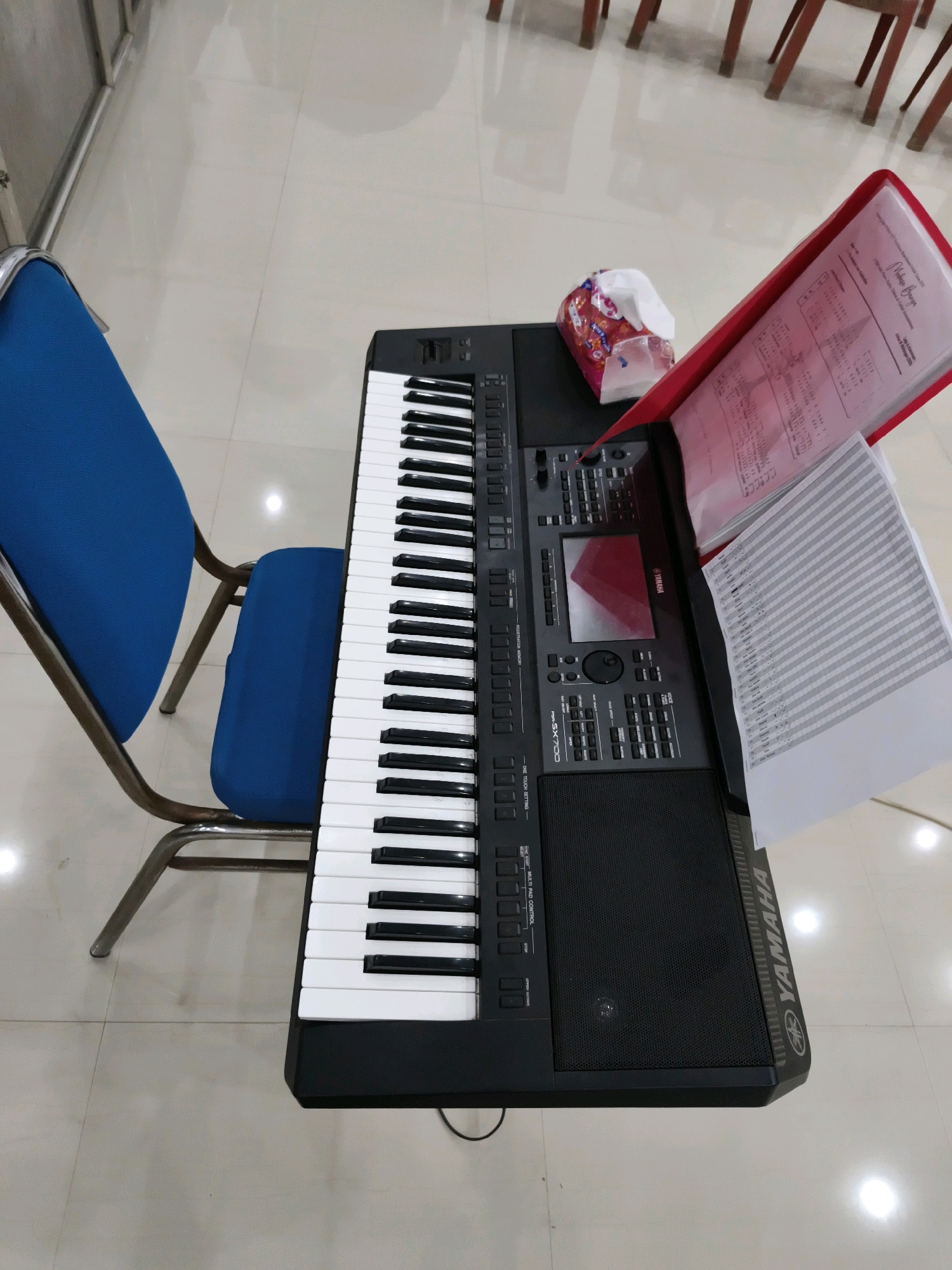 The image shows a Yamaha keyboard on a table with a blue chair next to it. There is a red folder with sheet music and a packet of tissues on top of the keyboard. The floor is tiled and appears to be in a well-lit room. This scene is located in Tombatu Dua, Indonesia. There are no people visible in the image.