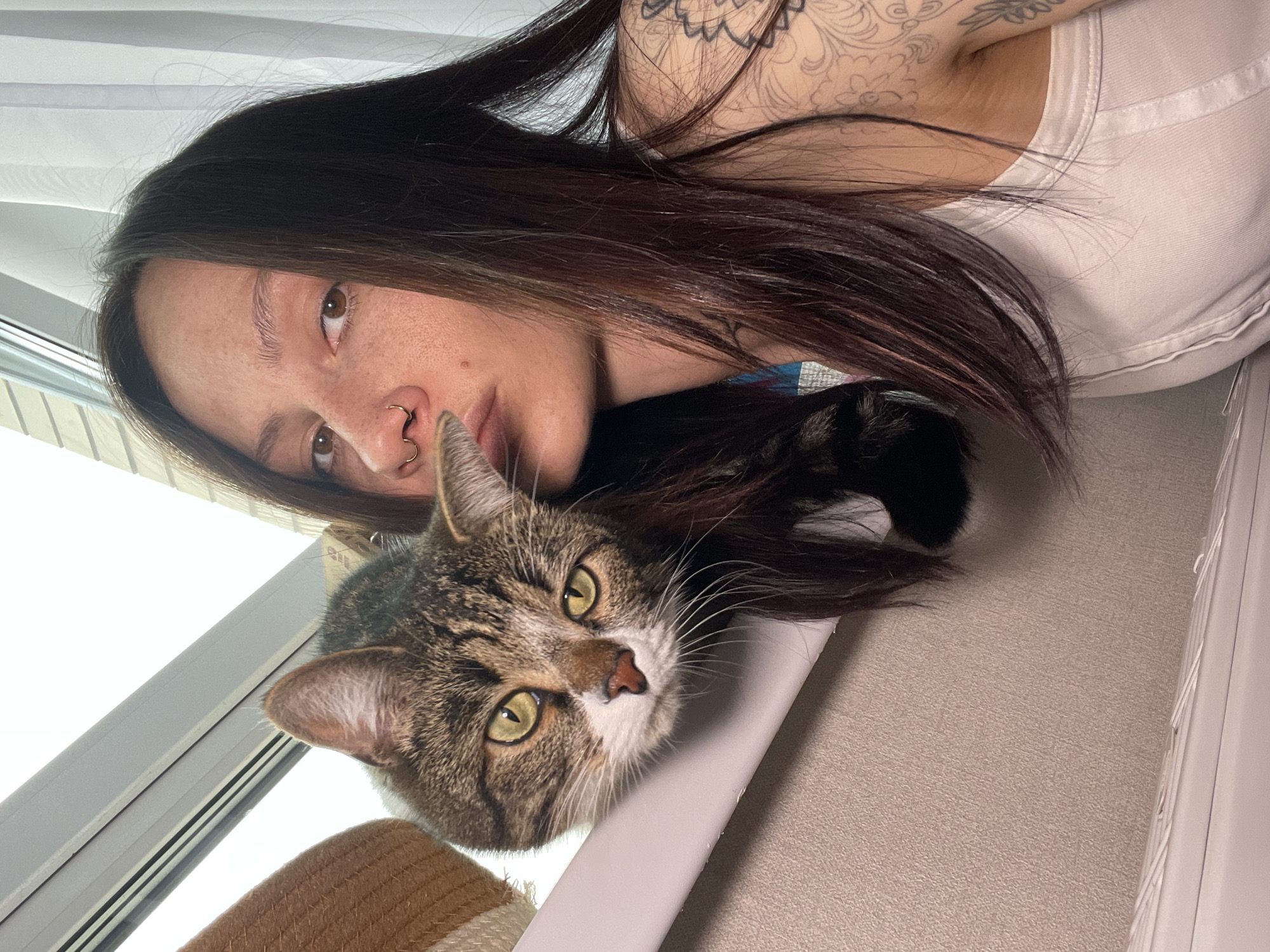 A young woman with dark hair and a white tank top lies beside a tabby cat. The woman has multiple piercings in her nose and a large tattoo on her upper arm. The cat has green eyes. They are positioned near a window with a beige wall and white trim. The exact location in Izhevsk, Russia, cannot be determined from the image.