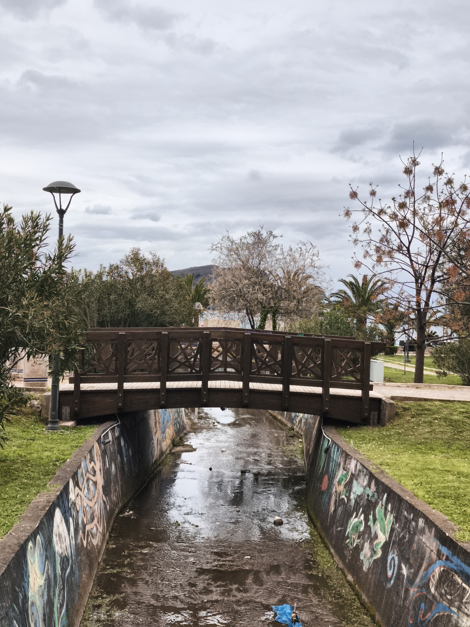 A medium shot depicts a canal bridged by a wooden footbridge within a park setting in Budva, Montenegro, under an overcast sky. The canal's concrete walls, heavily covered in graffiti, contain water with visible debris. Green grass borders the canal on both sides, leading to the bridge. The bridge's dark wooden construction features a decorative lattice pattern on its side rails. A lamppost stands adjacent to the canal. Trees and vegetation are visible beyond the bridge, with a mountain or hill discernible in the distance. The overall scene suggests an urban park area with signs of neglect.