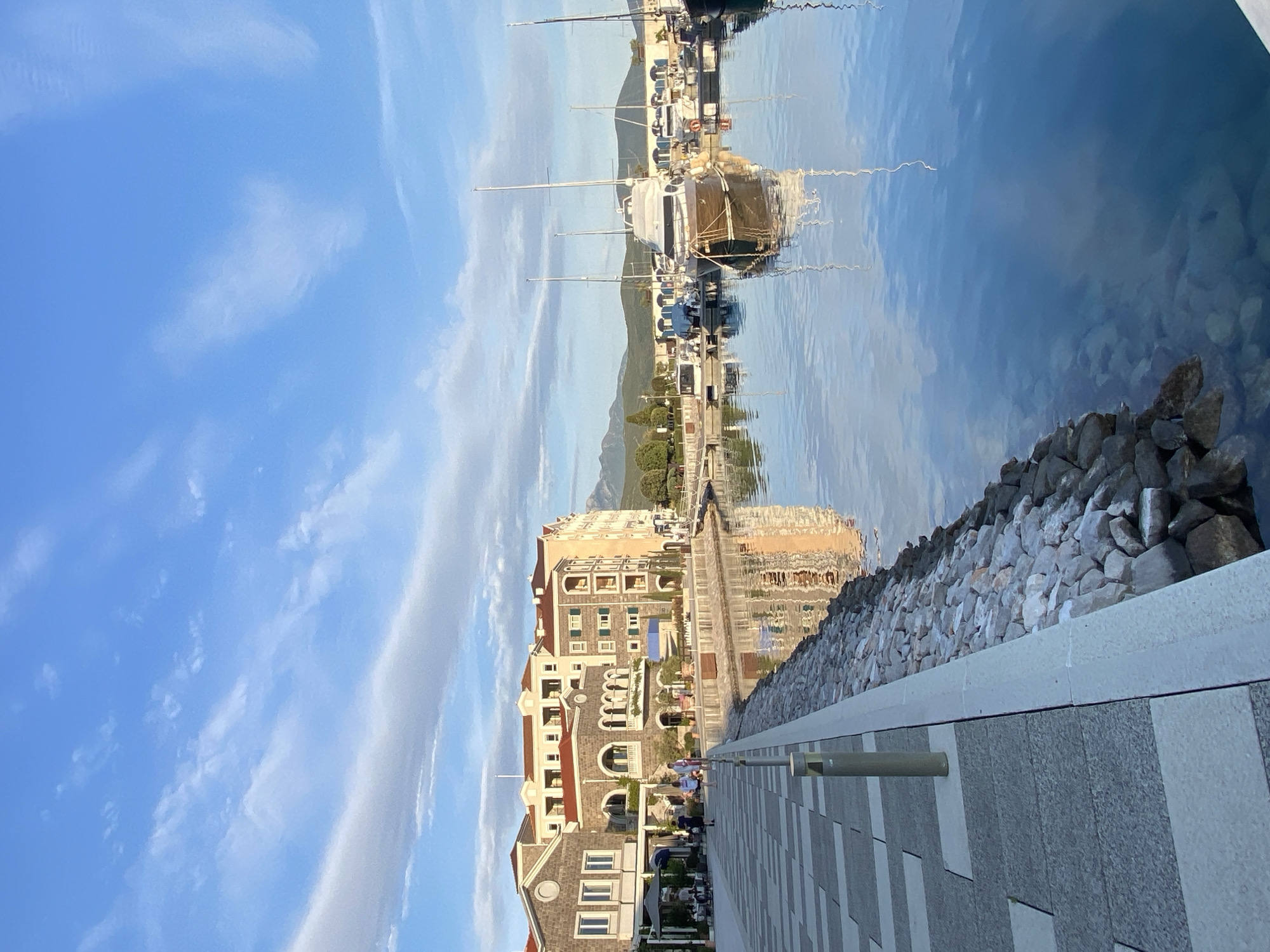The image captures a vibrant waterfront scene in Radovići, Montenegro, under a clear blue sky dotted with wispy white clouds. The perspective is from a paved promenade looking down a bustling marina.
On the left side of the frame, a series of multi-story buildings, likely hotels or apartments, line the waterfront. Their architecture features terracotta roofs, arched windows, and balconies, reflecting a Mediterranean style. A few people are visible strolling along the wide, patterned stone promenade or sitting at outdoor establishments near the buildings.
The right side of the image features the calm, reflective blue waters of the marina. Numerous boats and yachts of various sizes are docked, their masts and hulls casting rippled reflections on the water's surface. A low stone retaining wall separates the promenade from the water, with larger rocks visible below the waterline, suggesting a rocky seabed or shore. In the background, rolling green mountains provide a scenic backdrop to the bay.
The time of day appears to be bright daylight, possibly morning or late afternoon, given the clear conditions and soft illumination. The overall atmosphere is one of leisure and tranquility, typical of a coastal resort town. No discernible text is visible in the image.