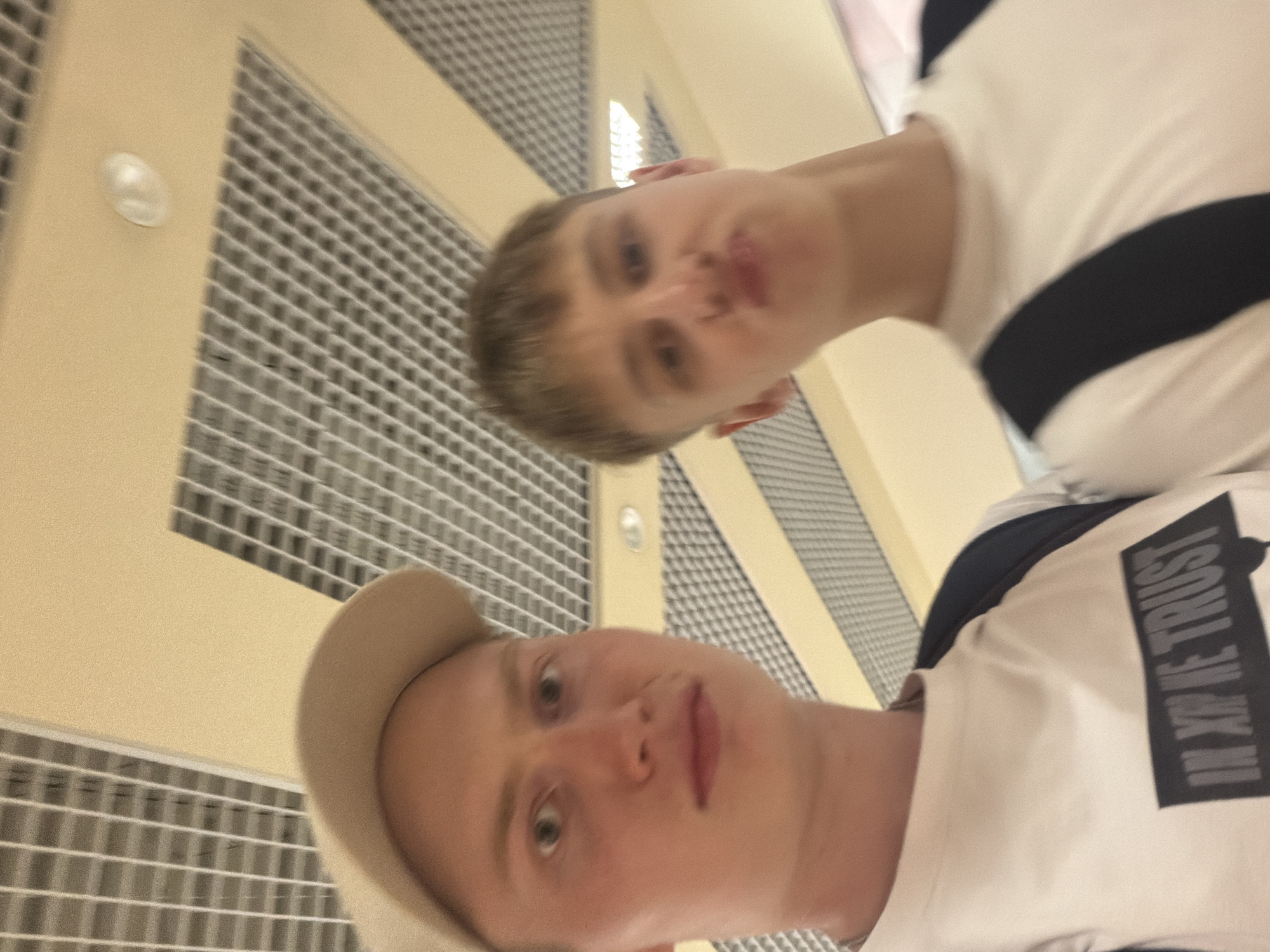 Two young men are posing for a selfie. The man on the left is wearing a beige baseball cap and a white t-shirt with the text "IN GOD WE TRUST" printed on it in black letters. The man on the right is wearing a white t-shirt and has his mouth puckered. They are standing in what appears to be an indoor space with a suspended ceiling that has a grid pattern. The lighting is bright and diffused. The background is out of focus, but shows a similar ceiling structure. Without more context, it is impossible to determine the specific location within Kyiv, Ukraine.