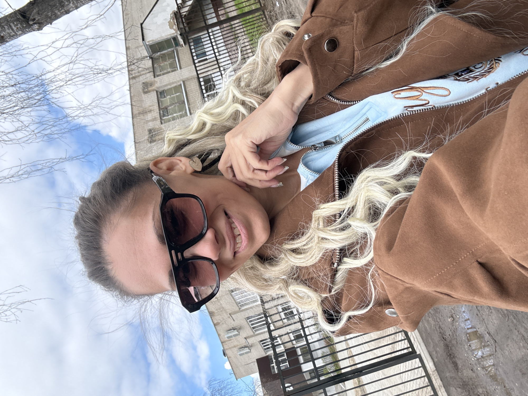 A woman with blonde hair and sunglasses poses for a selfie. She is wearing a brown jacket over a light blue zippered sweatshirt. In the background, there is an apartment building. The location is Rusanovo, Russia.