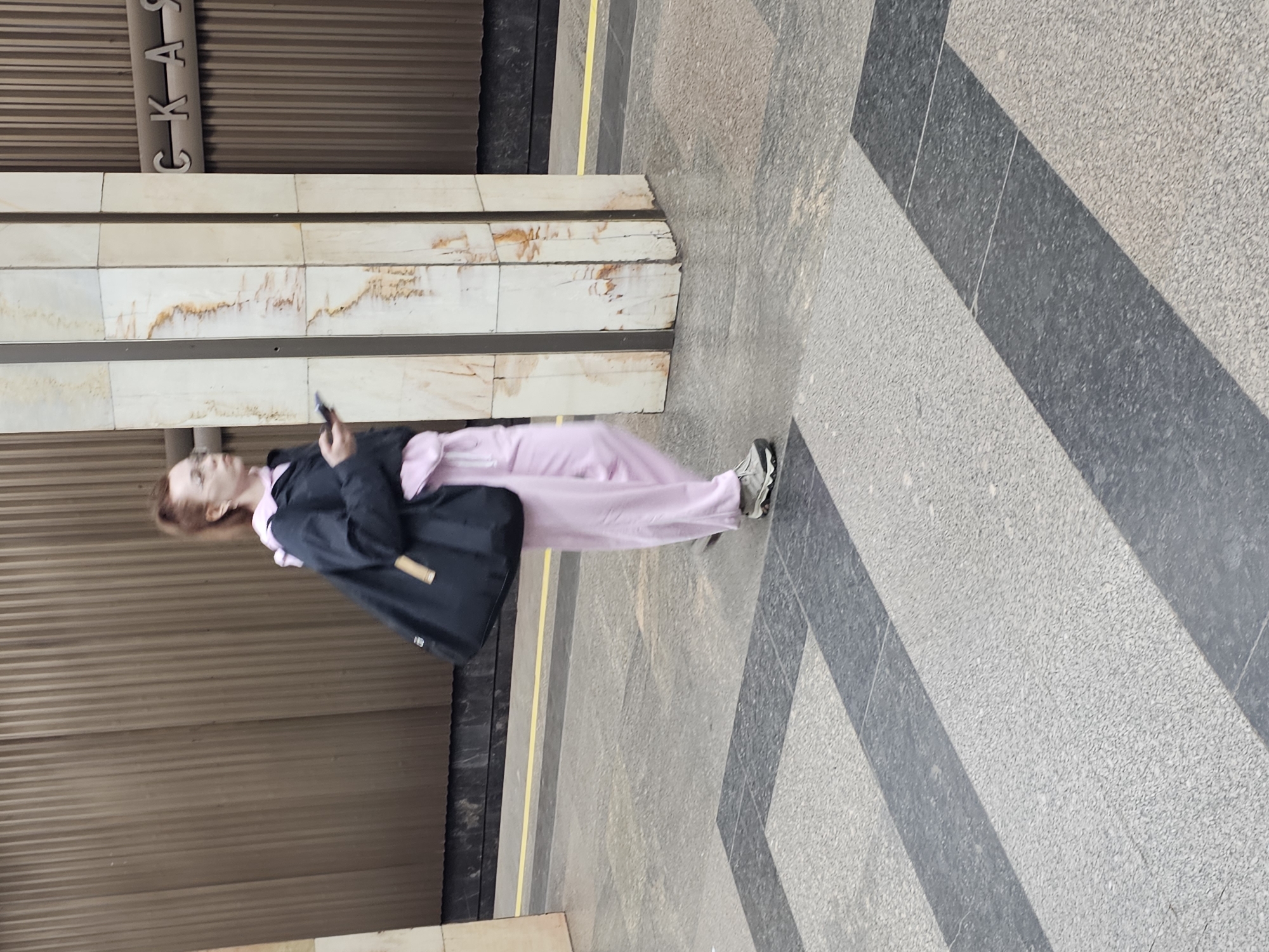 A woman is depicted standing on a subway platform, engrossed in her mobile phone. She has reddish-brown hair and is dressed in a light pink tracksuit, a dark jacket, and light-colored athletic shoes. A dark shoulder bag with a lighter handle is visible on her side.
She stands beside a rectangular support column, which is faced with light-colored marble or stone, showing natural brown patterns and some darker discolorations. The platform floor beneath her features a light grey speckled texture interspersed with darker grey geometric patterns, including diagonal stripes. A thin yellow line runs along the edge of the platform near the column.
The background wall is covered with horizontally corrugated brown metal panels. Above the column, partial Cyrillic lettering is visible, spelling out "СКАЯ" (SKAYA), which is a common suffix for Moscow Metro station names. The setting is clearly a subway station in Moscow, Russia, capturing a typical scene of urban transit waiting.