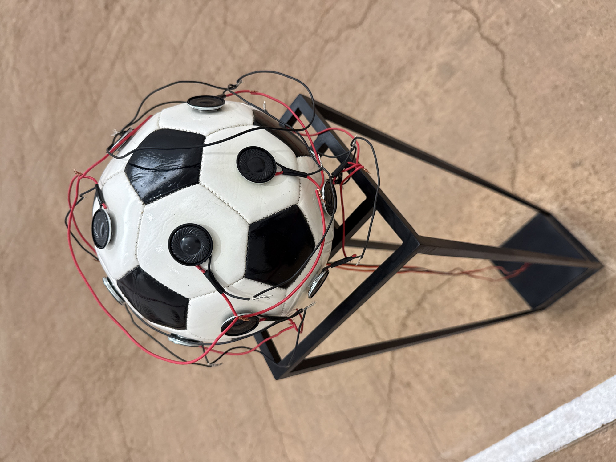 A white and black patterned soccer ball, typical of a standard size 5, is displayed centrally on a minimalist black metal stand. Numerous small, circular audio speakers, approximately 1-2 inches in diameter, are affixed to the surface of the soccer ball. These speakers are interconnected by a network of exposed red and black electrical wires, which crisscross the ball's panels. The wiring appears functional but not fully concealed, with some bare wire ends visible. The soccer ball rests securely within the square top frame of the open-sided stand, which has four vertical supports and a square base, all constructed from dark metal. The stand is positioned on a light brown or tan floor, possibly concrete, exhibiting subtle tonal variations and faint crack patterns. A partial white line, consistent with floor markings, is visible in the lower-left corner of the image. The scene is located in the city of Mexico City, Mexico. No individuals are present.