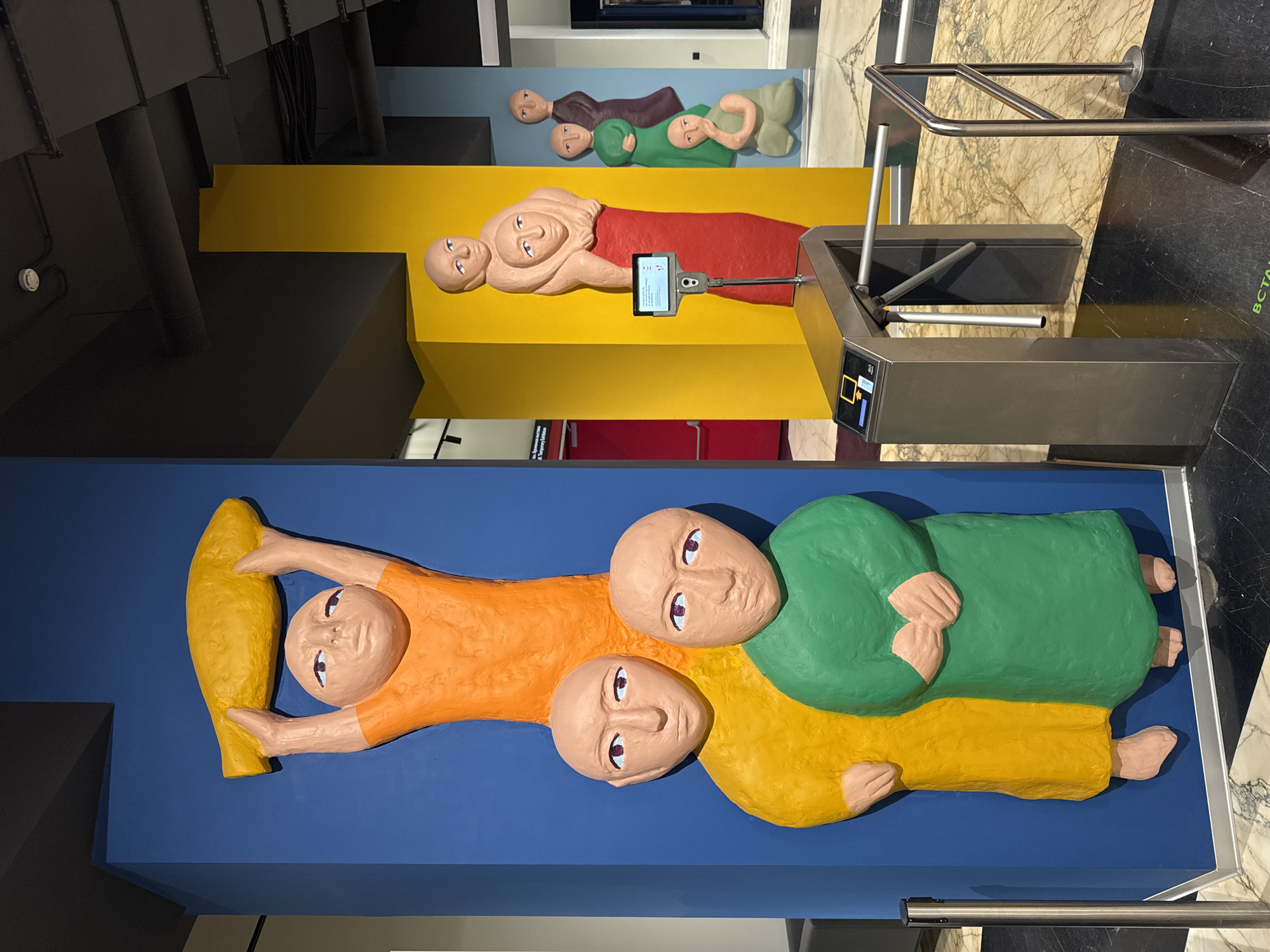 The image depicts an interior space, likely an entrance or exhibition area within an art museum or gallery, featuring colorful, stylized art installations alongside functional turnstiles.
On the left side of the scene, a vibrant blue wall displays three flattened, sculptural figures. An orange figure, with a bald head and simplified facial features, holds a large, yellow, fish-like object horizontally above its head. Below it, two intertwined figures lie horizontally: one in green with crossed arms and another in yellow. All figures share similar neutral or slightly melancholic expressions.
In the middle, a bright yellow platform or wall showcases a large, red, flattened figure with two smaller figures intertwined within its form. One small figure has its arms around the larger one's neck. A small digital tablet, mounted on a stand, is attached to the red figure, displaying text that is too small to be clearly read, likely providing information about the artwork.
Further to the right, a light blue wall features three more stylized figures: one in purple, one in green, and one in light green/beige, all lying horizontally and appearing to be stacked or intertwined.
To the right of the art installations, two modern, metallic turnstiles stand on a patterned floor. The floor transitions from a dark, possibly black marble, pattern near the turnstiles to a lighter, yellowish-brown marble pattern further back. One turnstile displays a screen with an icon, suggesting a card or ticket reader. On the dark floor, a partial green word, "BCTA", is visible, likely part of a Russian word or sign. The space is well-lit, characteristic of an indoor exhibition with artificial lighting.