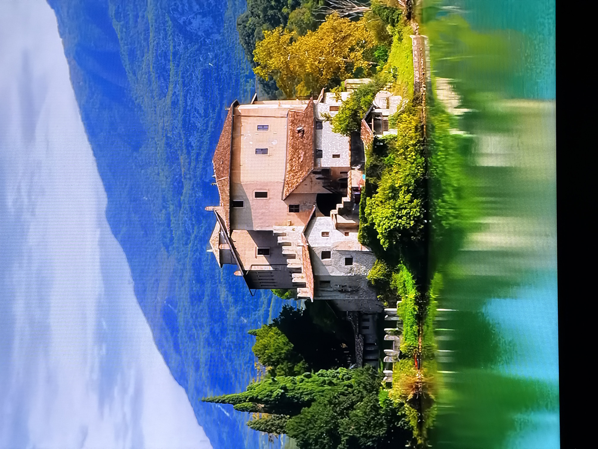 A picturesque scene unfolds featuring a stone castle situated on a small island in the middle of a serene, green lake. The castle, with its terracotta tiled roofs and crenellated battlements, is surrounded by lush trees and vegetation. In the background, majestic mountains rise, their slopes covered in dense forest, under a sky dotted with soft clouds. The water of the lake beautifully reflects the castle and the surrounding landscape, creating a mirror-like effect. The image does not depict the city of Chirchik, Uzbekistan.