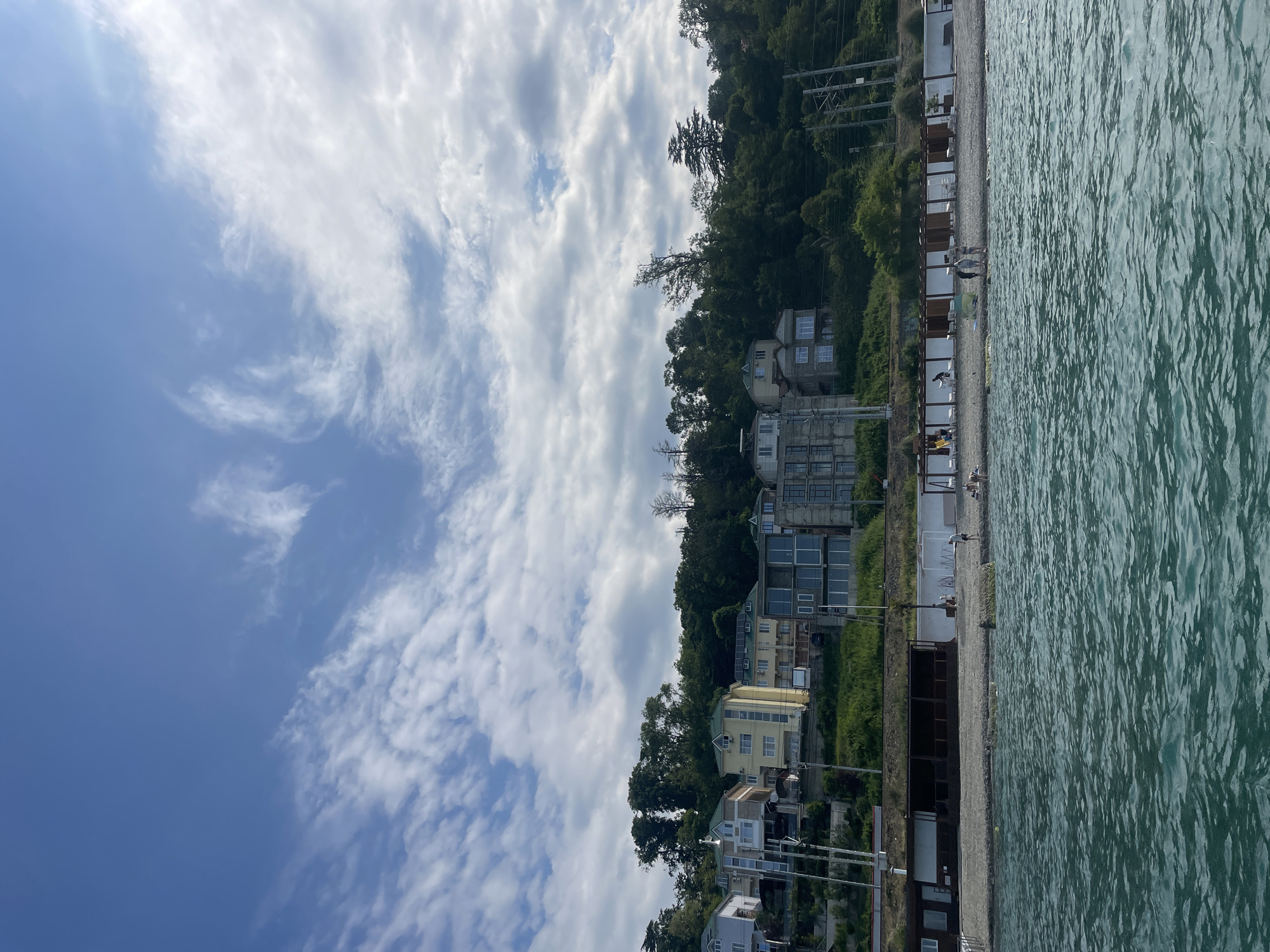The image depicts a coastal scene in Sochi, Russia, likely along the Black Sea. The foreground is dominated by the shimmering, greenish-blue water of the sea, showing gentle waves and ripples. Beyond the water is a pebble beach lined with a row of wooden structures that appear to be beach cabanas or seating areas. A few people are visible on the beach, some standing and others appearing to be walking or relaxing.
Behind the beach, a steep, tree-covered hillside rises, featuring a series of buildings built into the slope. These buildings vary in architectural style and color, with some appearing to be modern residences or hotels, characterized by large windows and balconies. Vegetation, primarily dense green trees, covers much of the hillside. The sky above is a bright blue with scattered white, wispy clouds, suggesting fair weather. Electrical poles and wires are visible along the hillside, indicating the presence of infrastructure.