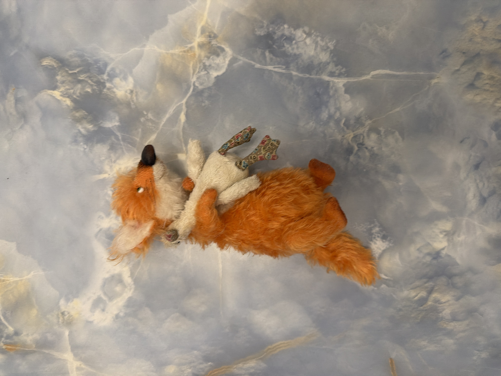 An orange plush fox toy is held by a white plush toy with patterned feet, set against a blue and white marbled background. The fox has a fluffy orange body, tail, and ears with white accents, and a black nose. The white toy has visible stitching and its feet are adorned with a floral pattern. The marbled background exhibits swirling patterns of light blue, white, and hints of yellow.