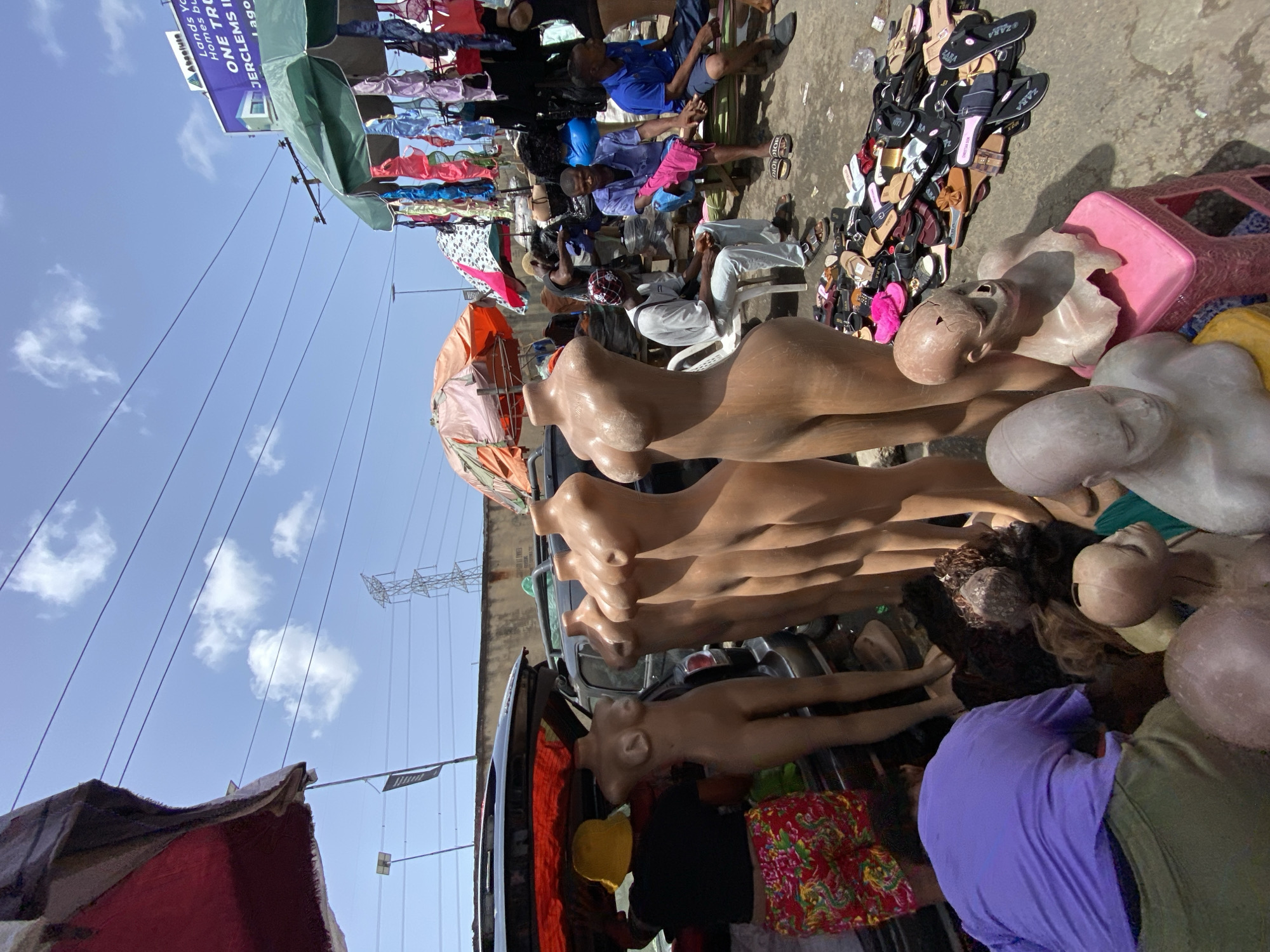 The image shows a bustling street scene in Lekki, Nigeria, with several vendors and shoppers. Multiple mannequins are piled up, some with damaged heads, along with wigs, clothing, sandals, and plastic stools, possibly for sale. People are seated in the background, some appearing to be engaged in conversation. A sign in the background advertises real estate services, stating "Lands Yo Homes bu ONE TRO JERCLEMS I Lago." Overheads are makeshift canopies providing shade. Power lines are visible against a blue sky.