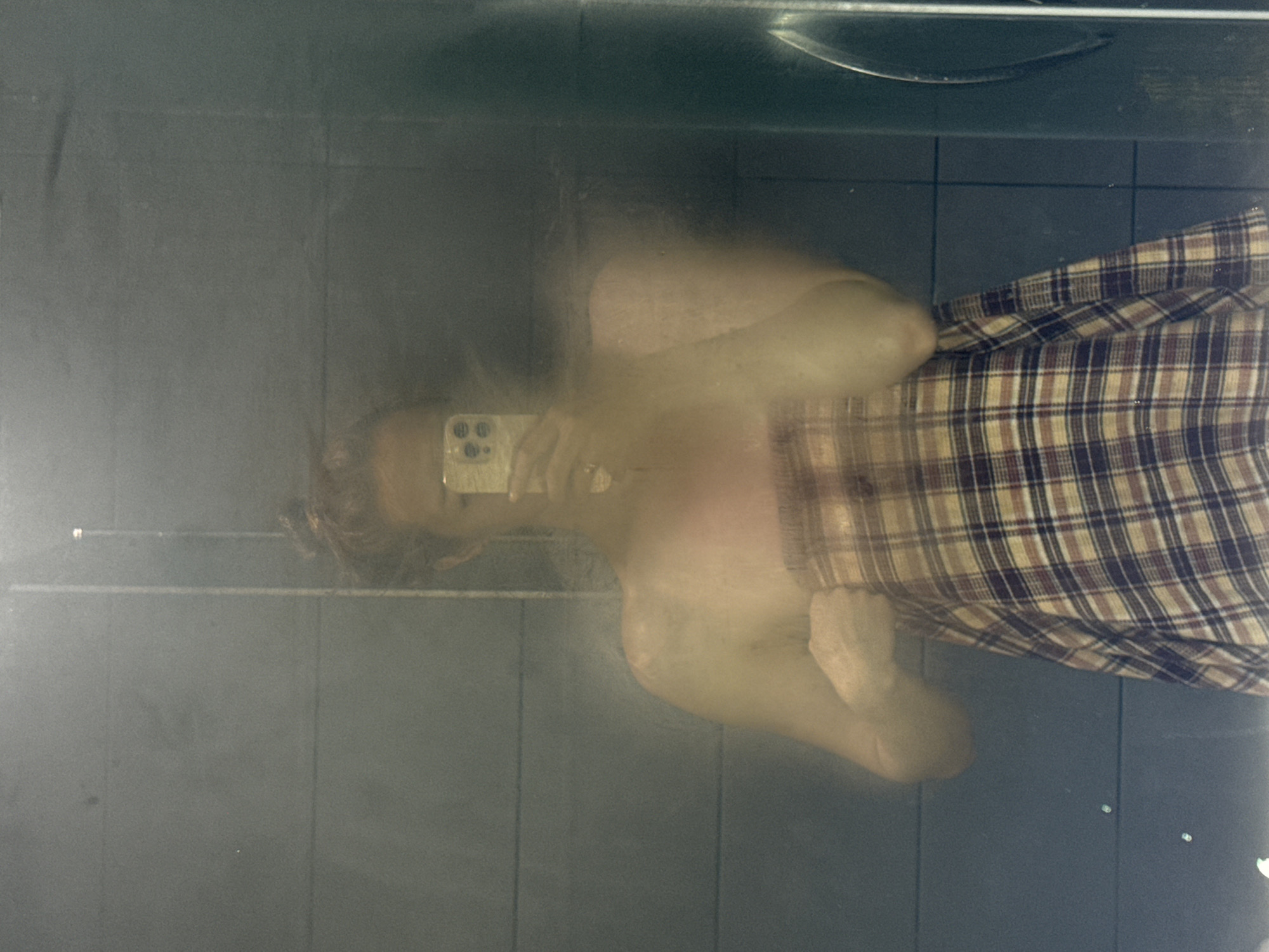 A blurred image shows a reflection of a person in a mirror, appearing to be in a bathroom setting, indicated by the tiled walls and what appears to be a sink or shower fixture at the top of the frame. The person is holding a cell phone, possibly taking the photo. The individual is partially wrapped in a plaid blanket or towel. The location of the image is claimed to be Chaikovsky, Russia, but there is nothing in the image to specifically confirm this.