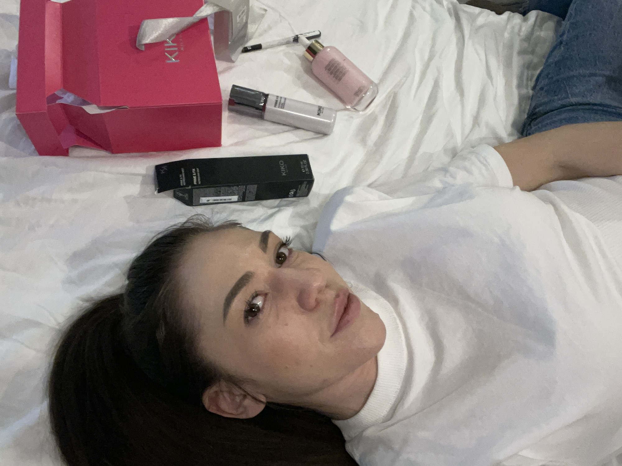 An indoor scene in Sofia, Bulgaria, depicts a female individual lying supine on a bed with white, crumpled sheets. The woman, appearing to be in her 20s or 30s, has dark hair pulled back, brown eyes, and is wearing a white long-sleeved top with a ribbed collar and blue jeans. Her head is angled slightly towards the camera, with her gaze directed forward. To her right, a black rectangular box labeled "KIKO MILANO" and "PRIME & FIX" is visible. Adjacent to the box are a light grey pump bottle and a pink cylindrical bottle with a gold-colored top, both also displaying "KIKO MILANO" branding. A thin, dark pen-like object lies near the bottles. Further to the right, an open hot pink gift box, branded "KIKO MILANO," is present, with white tissue paper visible inside. The environment suggests a personal space, likely a bedroom.