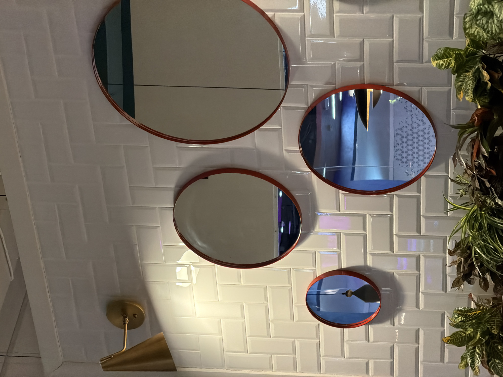 An indoor scene showcases a white tiled wall or ceiling arranged in a herringbone pattern, featuring a glossy finish that reflects ambient light. Four circular mirrors of varying sizes, each with a reddish-orange frame, are mounted on this tiled surface.
The largest mirror, positioned at the top left, reflects a plain, light-colored surface with a dark horizontal line. The second largest mirror, located at the top right, shows a reflection of an interior space dominated by blue and purple hues, vertical structural elements, and a subtle patterned surface on its right side. The third mirror, centrally placed on the left, reflects a light-toned surface similar to the largest mirror, with a dark vertical strip on its right edge. The smallest mirror, at the bottom right, reflects a blue-tinted area with a dark, conical object visible at its center, likely a reflection of a light fixture or decor.
To the far right, a vertical garden or plant arrangement featuring a variety of green and darker-leaved plants adds an organic element to the setting. In the bottom left corner, a brass-colored wall lamp with a conical shade is visible, mounted on a white, possibly angled, surface.
The overall impression is that of a modern and stylish interior space, likely a commercial establishment given the decor. No people or explicit activities are visible, presenting a static snapshot. The lighting suggests an indoor setting, possibly evening or artificial illumination. This scene is located in Izhevsk, Russia. No visible text is present in the image.