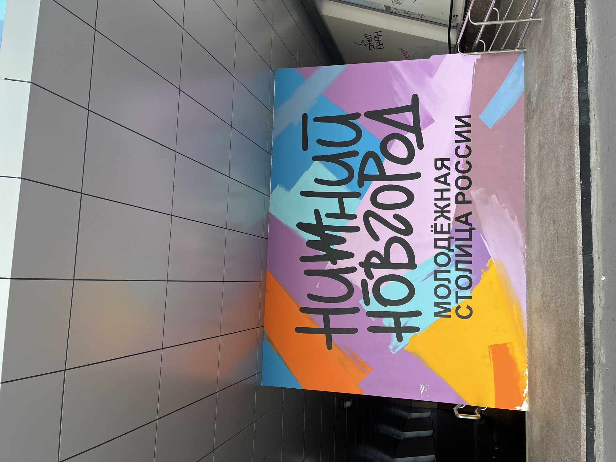 A large, vibrantly colored mural is displayed on a concrete wall under a modern, silver-paneled overhang. The mural features abstract splashes of orange, cyan, light blue, purple, and yellow. Prominently displayed in bold black lettering across the mural are the words "НИЖНИЙ НОВГОРОД" (Nizhny Novgorod), with smaller text below reading "МОЛОДЁЖНАЯ СТОЛИЦА РОССИИ" (Youth Capital of Russia). To the left of the mural, a silver door handle is visible on a dark entrance. To the right, a silver metal railing guides a ramp downwards, with graffiti visible on a concrete pillar in the background. The ground in front of the mural is a textured, gray concrete surface.