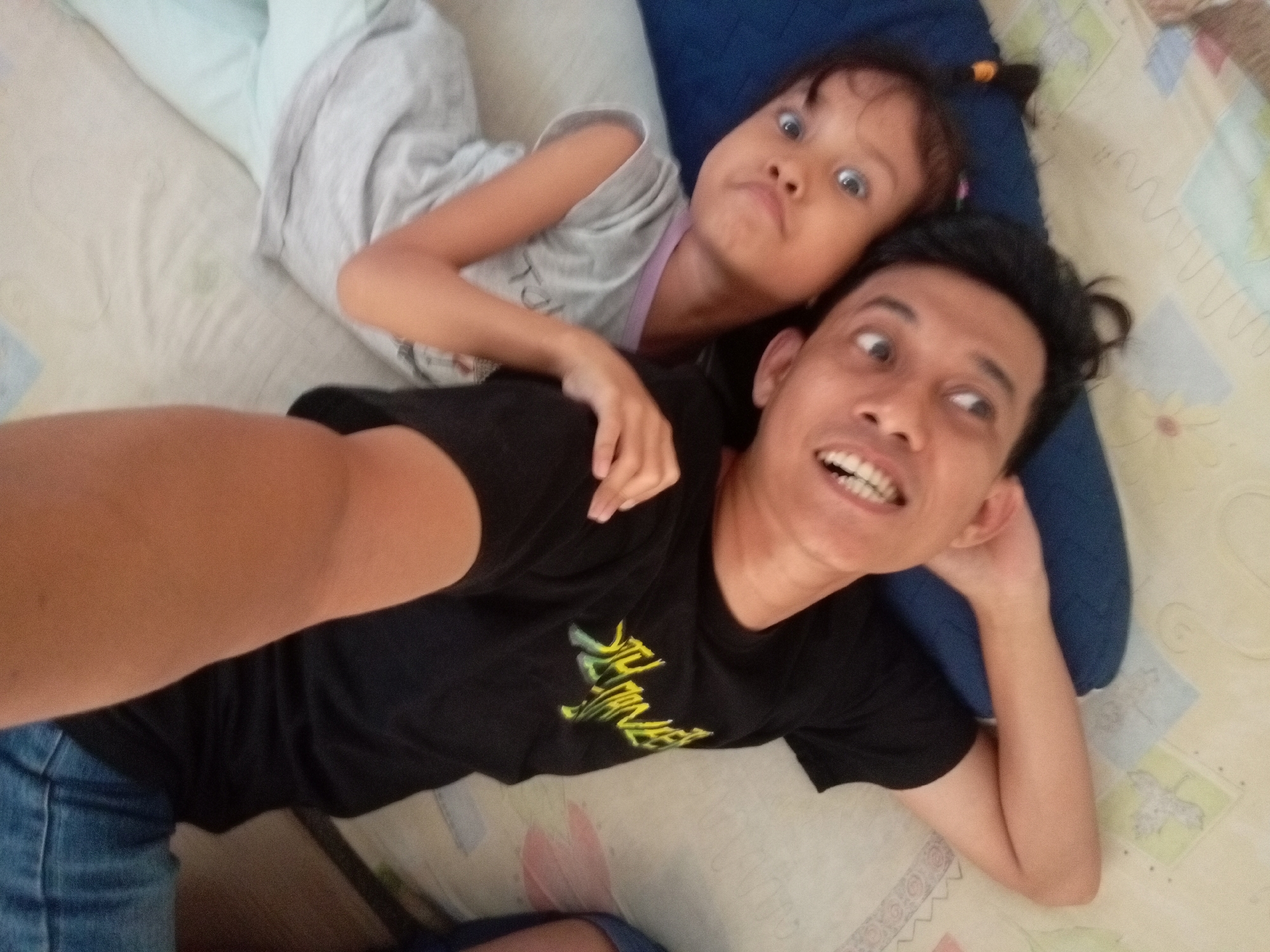 In this selfie taken from a high angle, a man and a young girl are lying on a bed, both looking towards the camera with exaggerated expressions. The man is in the foreground, wearing a black t-shirt with a yellow graphic design and blue jeans. He has a wide, open-mouthed smile, revealing his teeth, and his eyes are wide with a surprised look. The girl is positioned behind and slightly above him, resting her head on his shoulder. She's wearing a light gray t-shirt with a pink trim. Her eyes are also wide open, and her mouth is slightly ajar in a playful, surprised expression. A small orange hair tie is visible in her dark hair. They appear to be in a relaxed, indoor setting, likely a bedroom, with a patterned bedspread and a dark blue pillow visible. The lighting suggests it could be daytime, and the overall mood is one of lighthearted fun and playful interaction between the two individuals. The graphic on the man's shirt appears to be text, but it is not clearly legible.