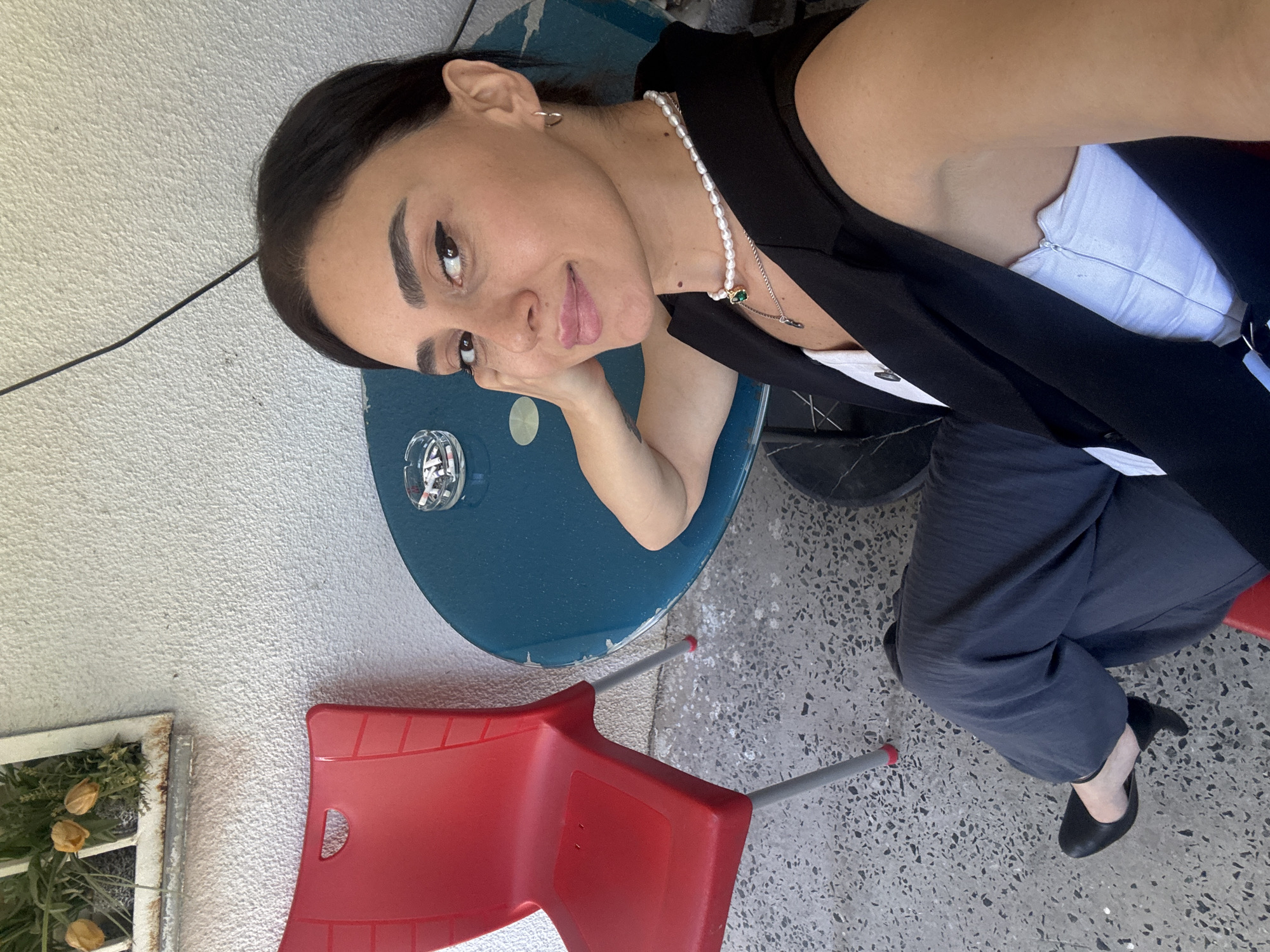 A woman with dark hair and a pearl necklace poses for a selfie. She is wearing a black vest over a white top and dark gray pants. She is sitting at a blue circular table with an ashtray containing cigarette butts. A red plastic chair is visible to her left, and a white textured wall with a window containing yellow flowers is behind her. The ground is a gray speckled concrete. The scene appears to be an outdoor seating area of a cafe or restaurant.