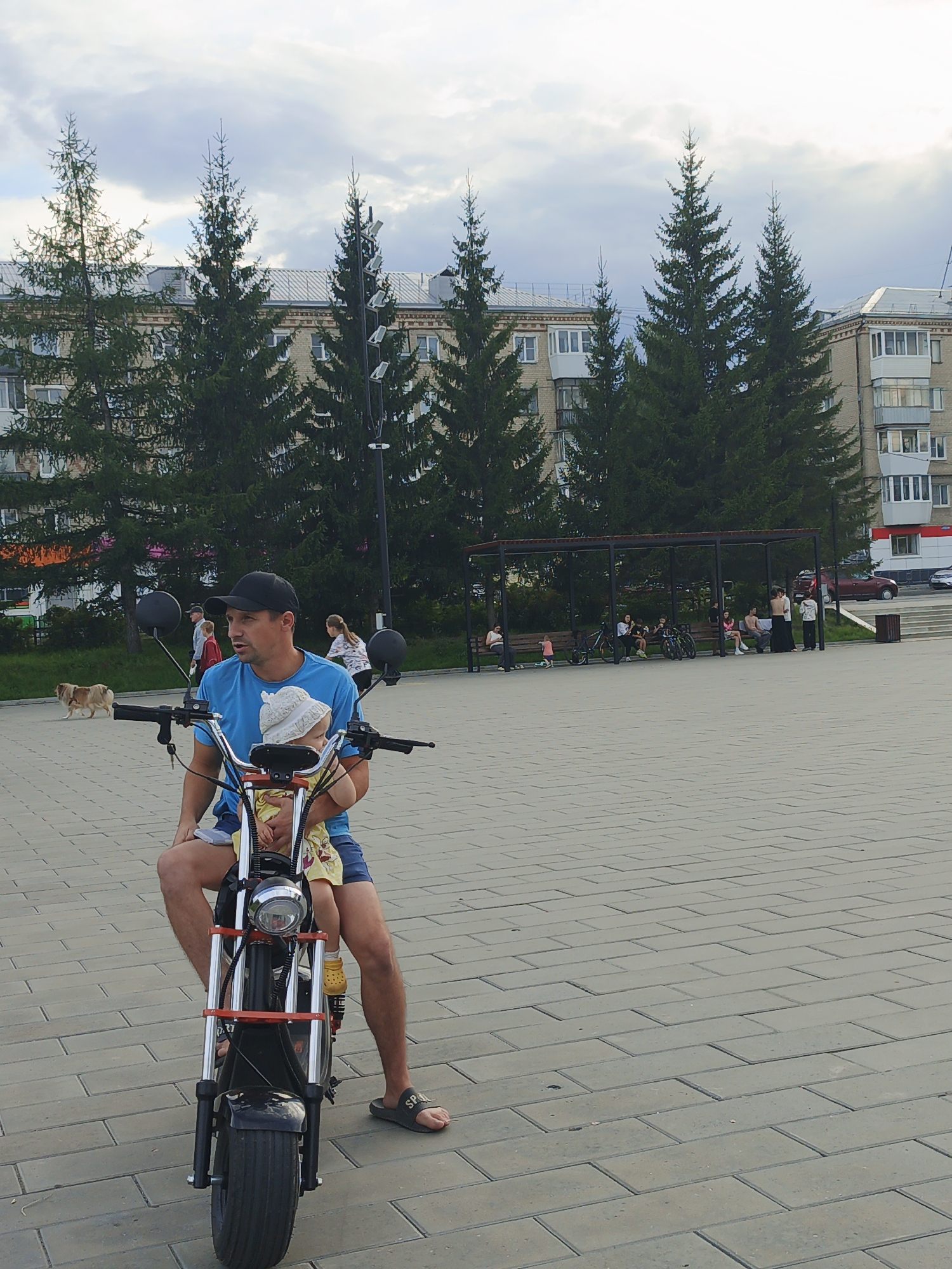 A man in a blue t-shirt and black baseball cap sits on an electric scooter in a paved plaza. He holds a young child in his lap, who is wearing a white hat and yellow dress. The man is looking to his right. In the background, several tall pine trees line the edge of the plaza, behind which are light-colored apartment buildings. A dog is visible on the left side of the frame, walking away from the scooter. To the right of the trees is a covered seating area with people sitting on benches. The ground is paved with rectangular bricks. The sky is overcast. This scene is consistent with a public space in the city of Zlatoust, Russia.