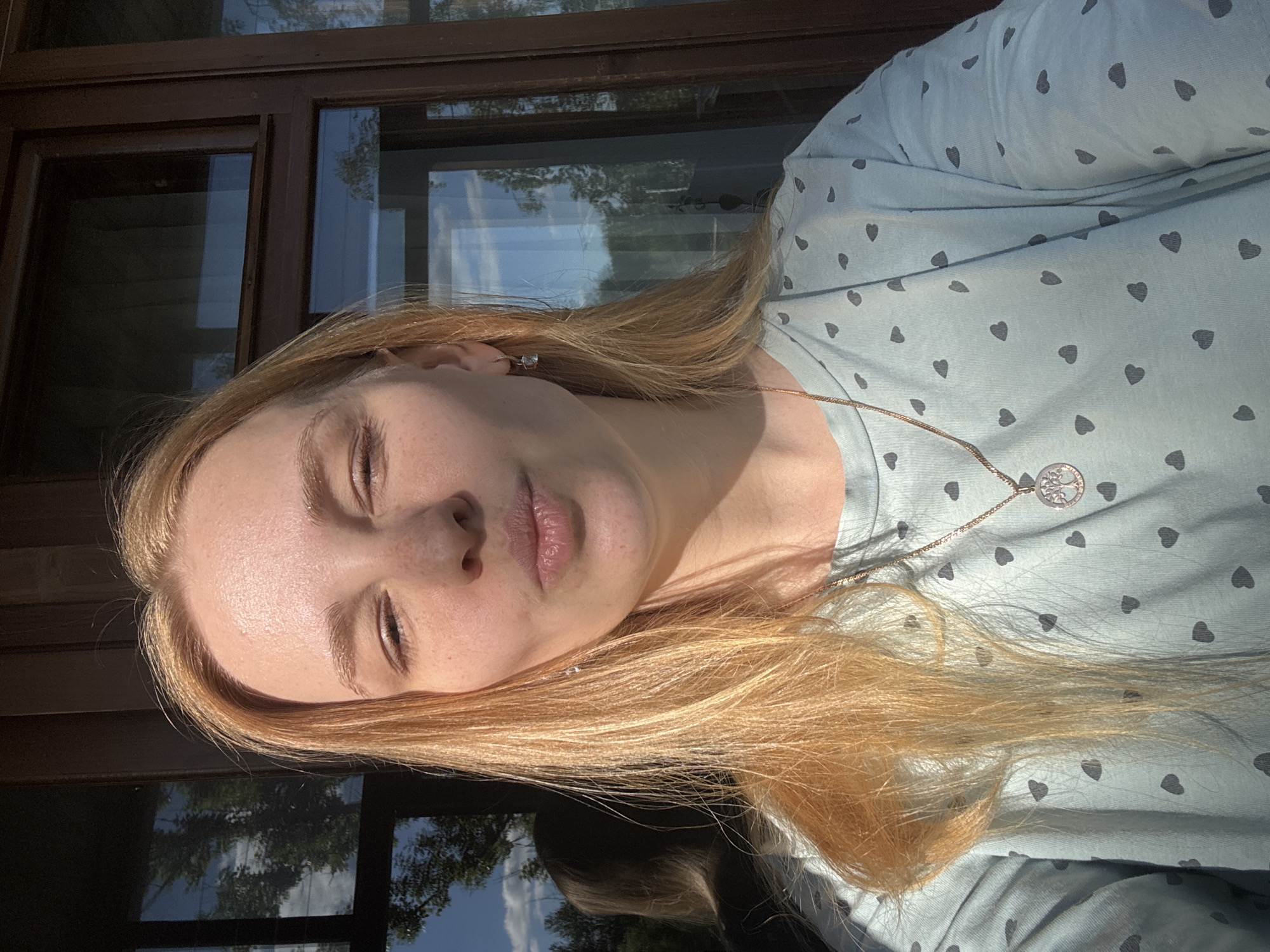 A young woman with long, flowing blonde hair, fair skin, and visible freckles is captured in a close-up portrait, rotated sideways. Her eyes are closed or heavily squinted, and her lips are slightly parted, suggesting a moment of relaxation or basking in sunlight. She wears a small, sparkly earring in her right ear and a delicate chain necklace featuring a circular, silver-colored pendant that appears to be a "Tree of Life" design. Her attire is a light blue or teal top adorned with numerous dark grey heart patterns.
The background reveals a dark-framed window, reflecting an outdoor scene. The reflections clearly show a bright blue sky, scattered white clouds, and the green foliage of trees. The strong, warm light illuminates the woman's face and hair, indicating a sunny day, likely during morning or late afternoon hours. The scene evokes a sense of peace and contentment. The location is Rusanovo, Russia. No discernible text is visible in the image.