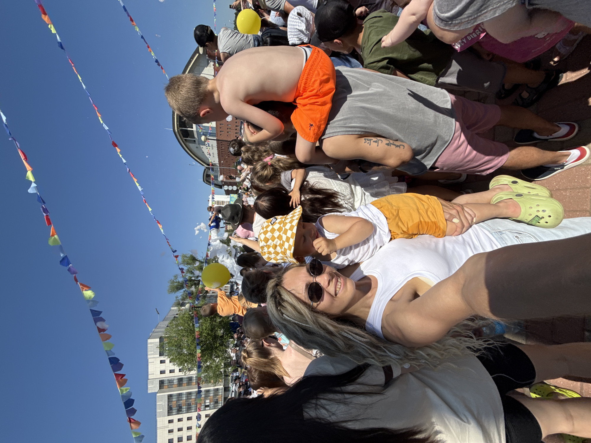 In Заволжье, Russia, on a sunny day, a crowd of people gathers outdoors, likely for an event. Colorful triangular flags are strung across the blue sky, adding a festive atmosphere. In the foreground, a woman with blonde dreadlocks and sunglasses smiles at the camera, holding a young child wearing a yellow and white checkered bucket hat and orange shorts. The child is being lifted by an adult. Further back, other children and adults are visible, some with balloons and bubbles. Buildings and trees form the background, suggesting an urban park or event space.
