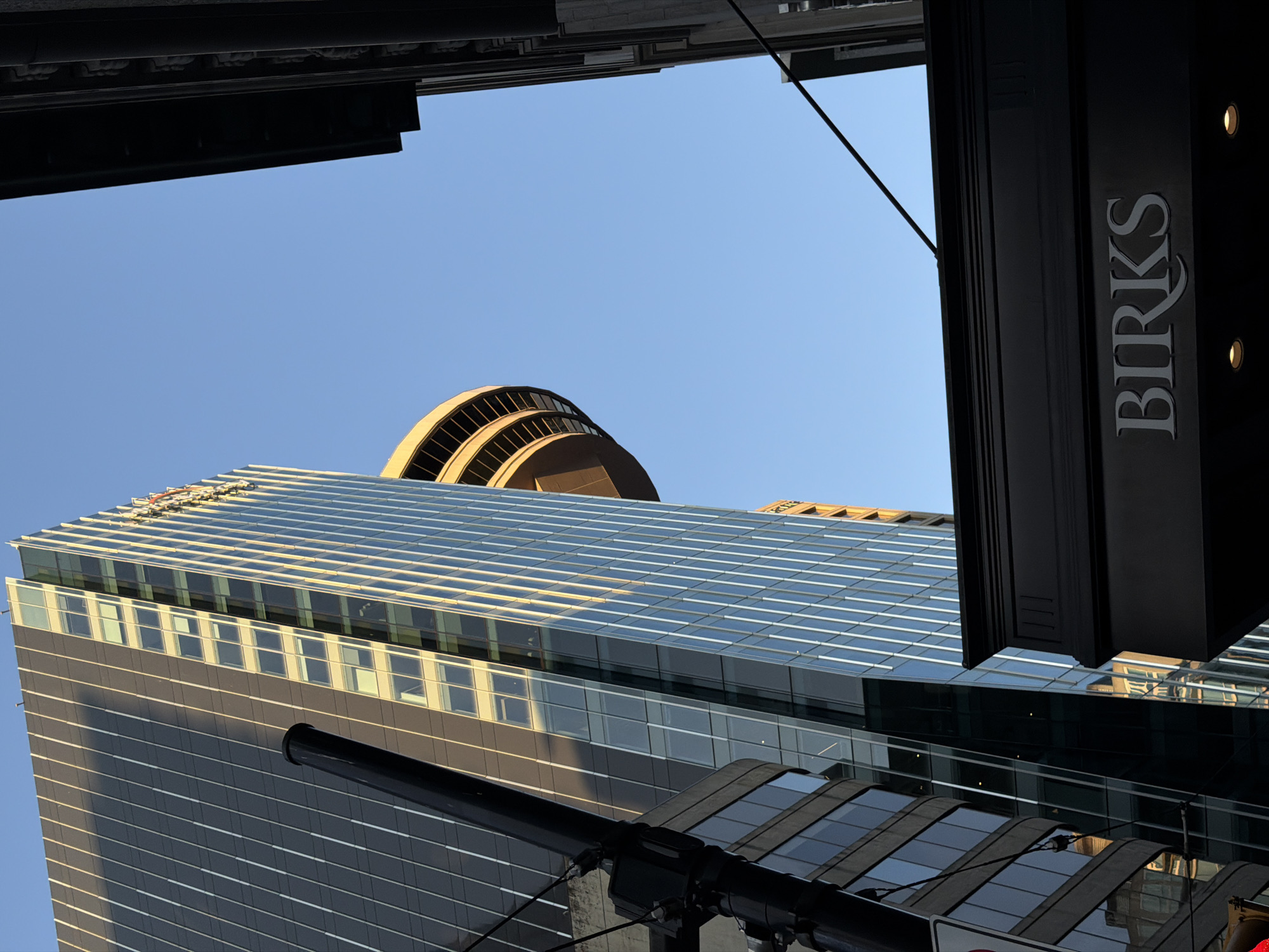 The image shows a view looking up at modern architecture in Vancouver, Canada. The primary focus is a tall, glass-facade skyscraper with vertical white lines, reflecting the clear blue sky and warm sunlight. To the left of this skyscraper, a portion of another building with darker glass and horizontal windows is visible, along with a black pole that appears to be part of a street light or utility fixture.
In the mid-ground, a distinctive circular, multi-story structure with a golden-yellow facade and visible windows protrudes from another building. To the far right, the dark facade of another building frames the scene, with ornate detailing visible along its top edge.
In the foreground, a dark, horizontal architectural element, likely part of a building's facade or an awning, spans the bottom portion of the image. The word "BIRKS" is prominently displayed in elegant white lettering on this dark element, identifying it as a Birks store, a well-known Canadian jewelry retailer. Two small, circular lights are visible beneath the "BIRKS" sign.
The composition suggests an urban setting with a mix of contemporary and possibly older architectural styles. The lighting indicates it is likely late afternoon or early morning, with the sun casting long shadows. There are no people visible in the frame. The overall impression is one of architectural grandeur against a clear sky.