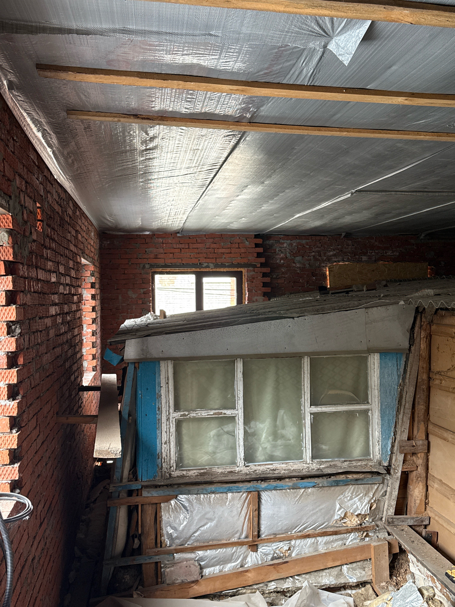 An indoor, low-angle, medium shot shows a construction site inside a building with exposed brick walls and unfinished ceilings. The ceiling is covered with reflective insulation material, supported by wooden beams. On the right side of the image, a dilapidated structure with a blue-painted wooden frame and a multi-paned window is visible. The window is covered with a translucent curtain. The area below the window is cluttered with building materials, including plastic sheeting and wooden planks. To the left, an unfinished brick wall extends into the background, revealing the rough texture of the bricks and mortar. A black corrugated conduit is visible near the bottom left corner. The overall scene depicts an interior space undergoing renovation or construction, characterized by unfinished elements and a generally unkempt appearance.