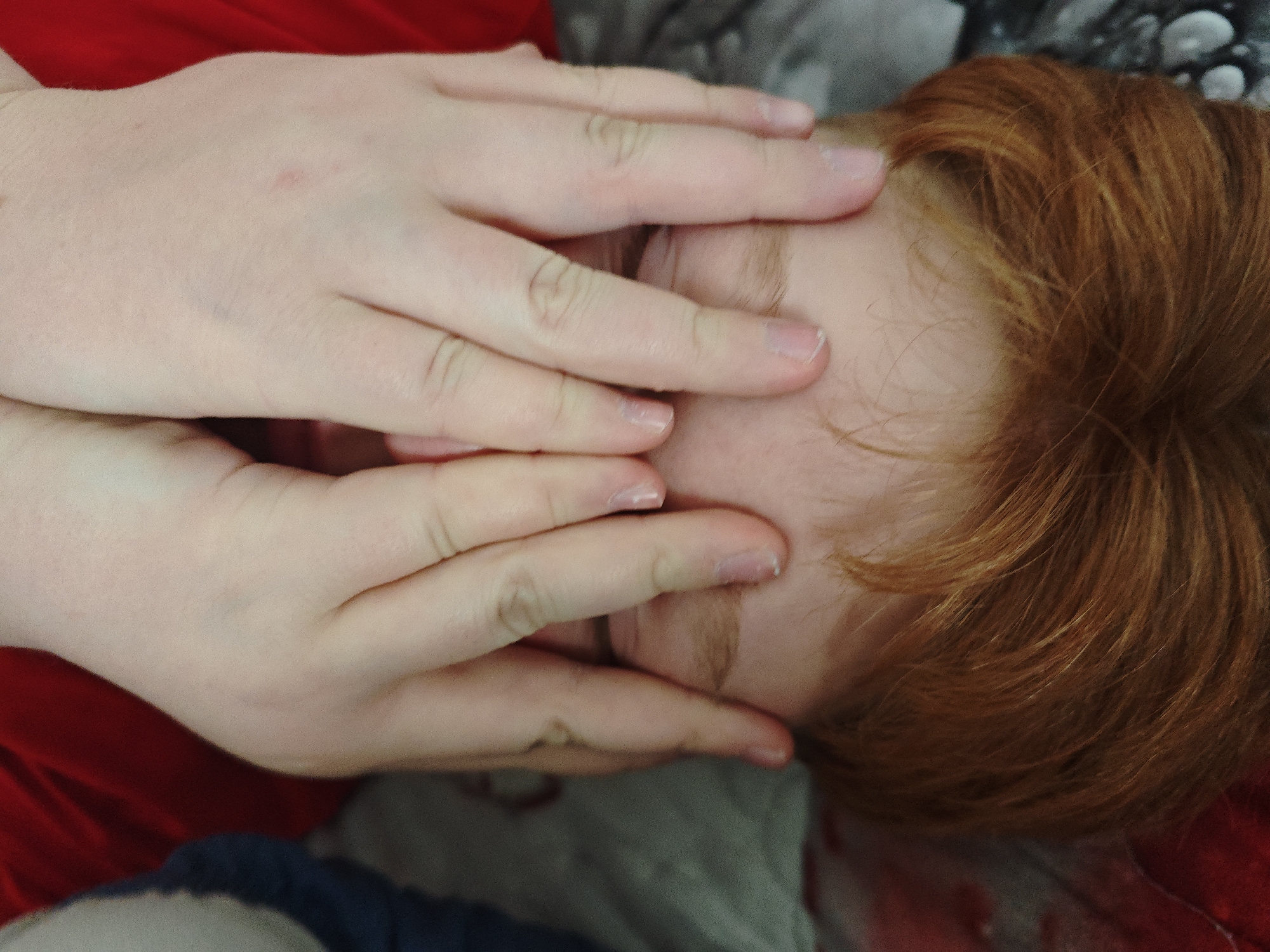 A person with red hair is lying down, face obscured by their hands. The person is wearing a red shirt. The background consists of a mottled gray and white surface. The location is not discernible from the image. No city is identifiable from the image.