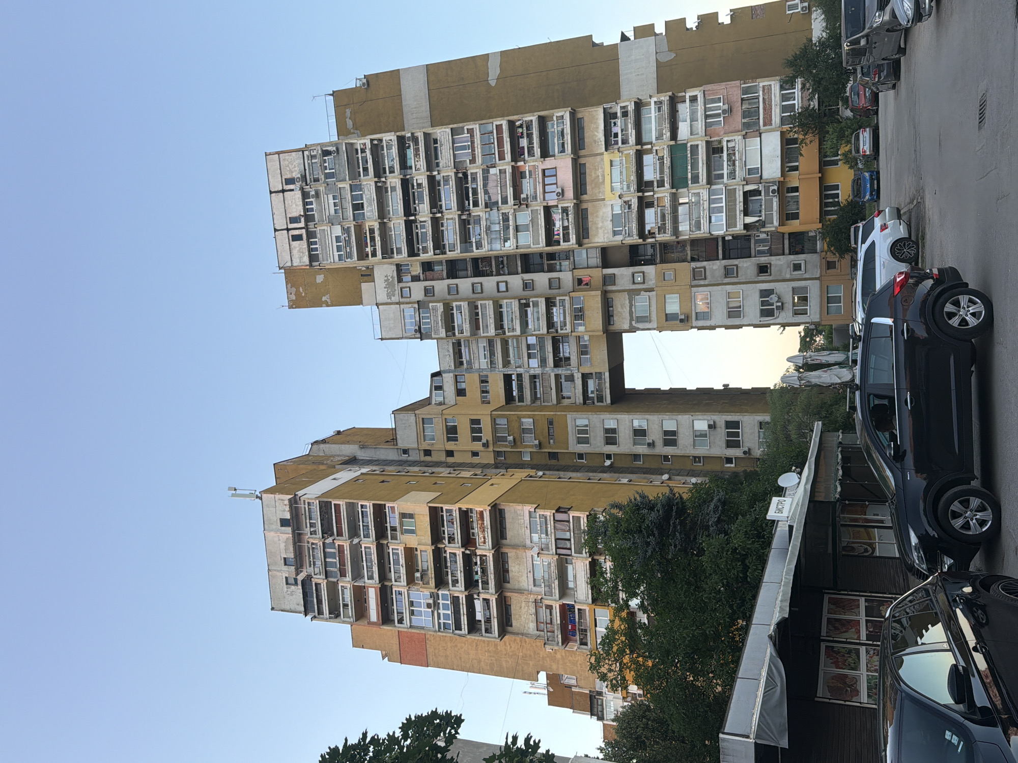 The image shows a brutalist residential building in Sofia, Bulgaria, characterized by its imposing H-shape, with two tall structures connected by a skybridge. The building, painted in shades of yellow and beige, features numerous balconies and windows, many with air conditioning units. The facade appears weathered, with some areas of peeling paint. In the foreground, several cars are parked on a paved area, and a small, low-rise commercial structure with awnings is visible on the left. Trees and greenery are present around the base of the building. The sky is clear and a light blue, suggesting it is either early morning or late afternoon. The overall impression is one of a functional, albeit dated, urban residential complex.