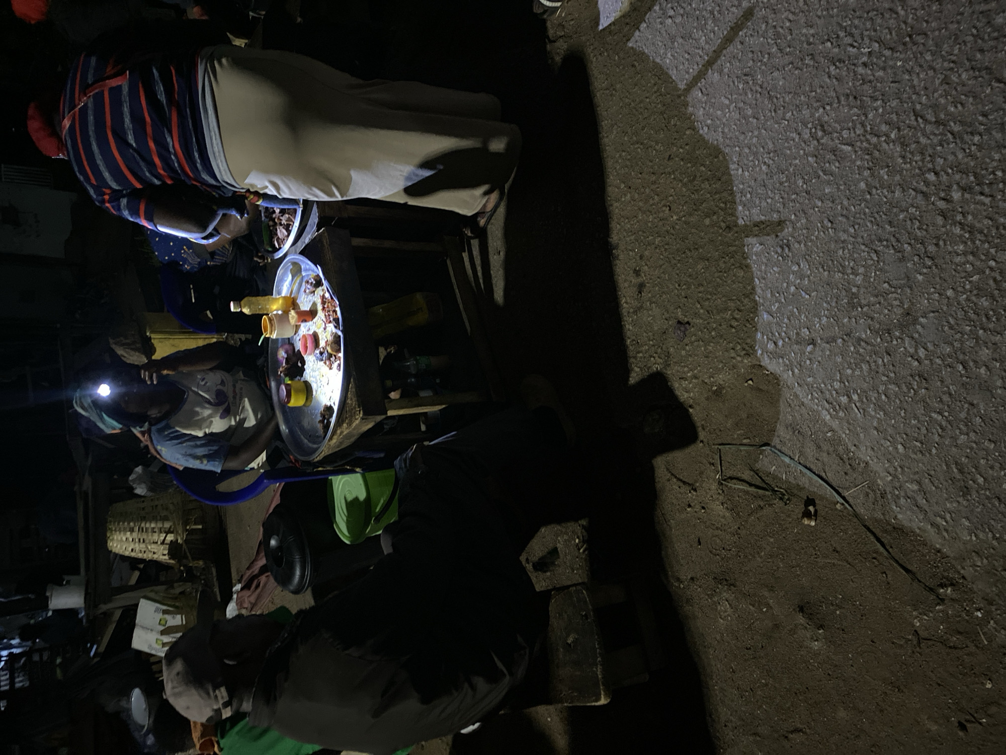 The image depicts a nighttime street food scene in Bukuru, Nigeria. Two individuals are seen preparing and serving food from a makeshift stall.
One person wears a headlamp and a light blue patterned headscarf and is seated on a blue plastic chair facing the food preparation area.
Another individual wears a striped shirt and is standing beside the food preparation area, apparently interacting with the food or the seated person.
A circular metal tray atop a small wooden table holds a variety of food items, including what appear to be fruits, vegetables, and sauces in bottles or containers.
The surrounding environment appears dark and somewhat cluttered with various items, including a green plastic bucket, a woven basket, and boxes. The ground is visible on the right side of the image, appearing to be a combination of dirt and pavement. Shadows cast by the lighting suggest a single light source in the food preparation area.