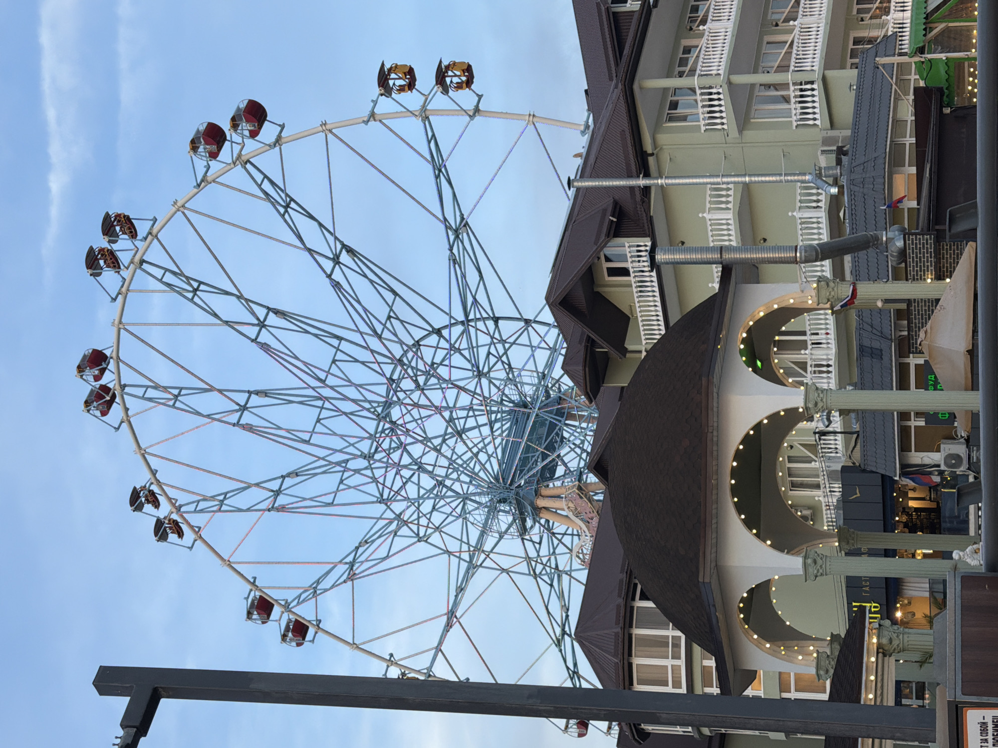 The image shows a large Ferris wheel against a partly cloudy sky. The Ferris wheel has several red cabins. In the foreground, there are several buildings with brown roofs, white balconies, and green walls. The buildings appear to be part of a resort or hotel complex. There are also decorative arches with string lights. The scene is likely located in Sochi, Russia, a popular resort city on the Black Sea coast, known for its amusement parks and tourist attractions. There are no people visible in the image.
