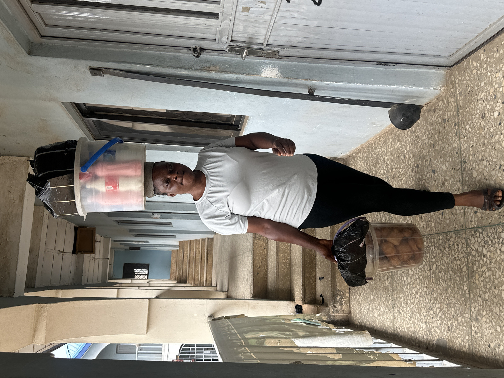 A woman walks down a covered walkway in Jos, Nigeria. She is balancing a large, clear plastic bucket containing bottles of liquid and black plastic bags on her head. She is also carrying another clear plastic bucket in her right hand, filled with fried dough balls, covered with a black plastic bag. The woman is wearing a light gray t-shirt and black leggings, and brown sandals. The walkway has a tiled floor and light blue walls. To the left of the walkway, there is a railing and a section of a building with a blue roof. Stairs are visible further down the walkway. To the right, there is a metal door.
