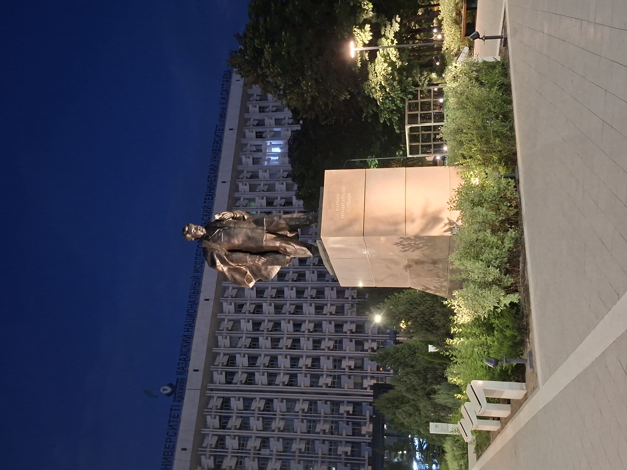 The image depicts the monument of Kanysh Satbayev, a prominent Soviet geologist and founder of the Soviet school of mining geology, located in front of the Kazakh National Research Technical University named after K. I. Satbayev in Almaty, Kazakhstan. The monument is a bronze statue of Satbayev standing on a stone pedestal. The university building, a large, multi-story structure with a repetitive architectural design, is visible in the background. The scene is illuminated by artificial lights, suggesting it is nighttime or dusk. The foreground features a paved walkway and well-maintained greenery, including bushes and trees. Several modern-style light fixtures are integrated into the landscaping. The overall impression is that of a well-lit public space in front of an educational institution.