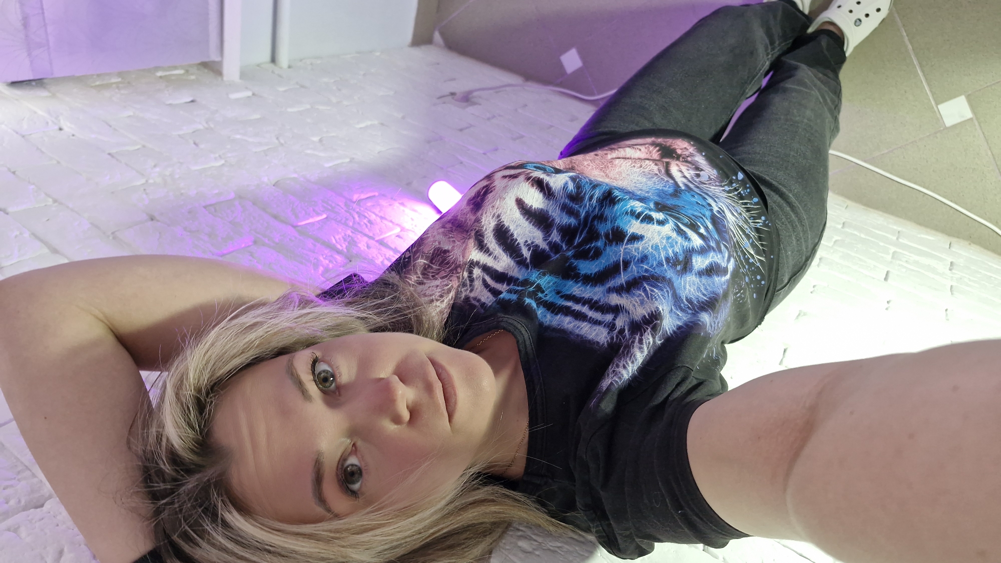 A woman is captured in a self-portrait, lying on her back and looking directly at the camera with a neutral expression. She has blonde hair and wears a black t-shirt adorned with a striking, multi-colored tiger head graphic in shades of blue, pink, and white. Her lower body is dressed in dark gray jeans and white Crocs-style shoes.
The setting appears to be indoors, in Otchor, Russia, characterized by a textured white surface, possibly a floor or wall, resembling painted brickwork. The scene is lit by artificial colored lights, with a distinct purple/pink glow emanating from a light source near her left arm, and a cooler, greenish-white light visible further in the background. A thin white cable is present on the floor. The controlled lighting suggests an evening or an intentionally designed indoor environment, possibly a studio or a room with decorative illumination. No discernible text is visible within the image.