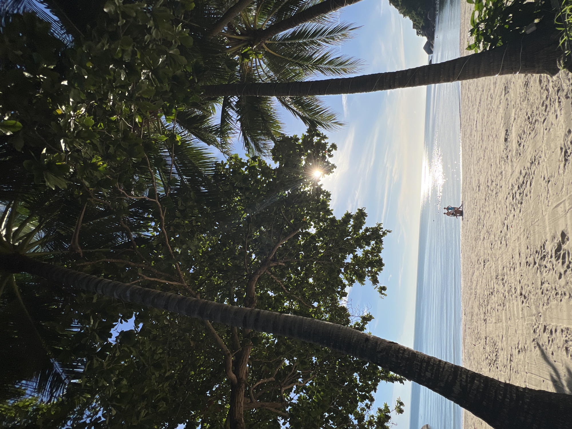 This is a wide-angle, eye-level shot of a tropical beach in Baan Chaweng Noi, Thailand. The foreground is dominated by the trunks and foliage of several palm trees, framing a view of the ocean and beach. The sand is light-colored and appears to have footprints scattered across it. Two individuals, one appearing to be in blue shorts and the other in light-colored shorts, are visible near the water's edge. The ocean is calm with gentle waves and reflects the bright sun. The sky is a clear blue with some wispy white clouds. The sun is shining through the palm fronds, creating a lens flare effect. In the distance, a lush green hillside with large rock formations is visible on the right side of the frame. The overall impression is a serene beach scene.