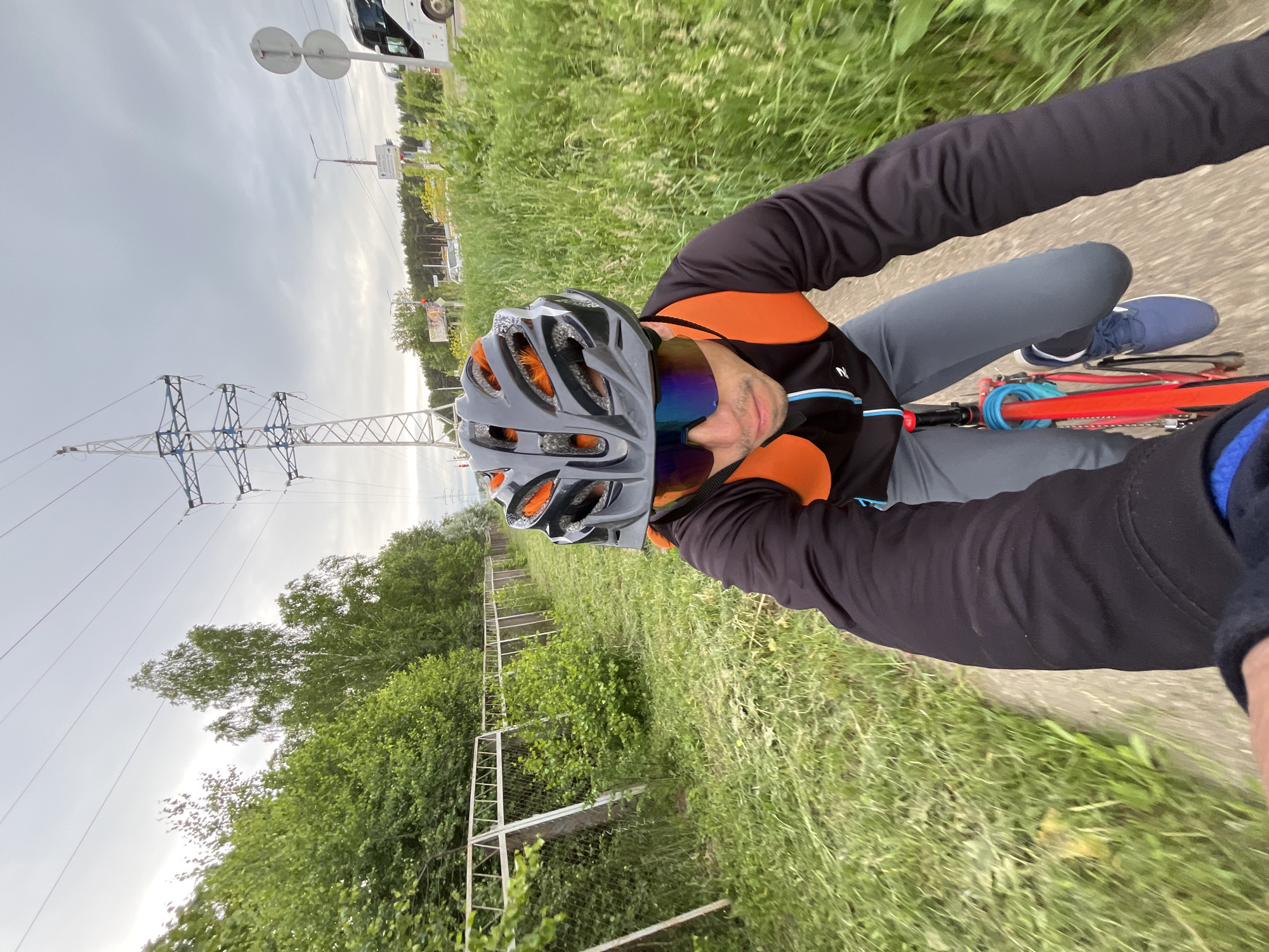 A first-person perspective, low-angle shot captures a cyclist riding on a path in Chaikovsky, Russia, under a cloudy sky. The cyclist wears a black and orange helmet, mirrored sunglasses, a black long-sleeved jacket with an orange vest, and grey pants. The cyclist's hands are visible, one holding the handlebars of a red bicycle. The path is bordered by tall green grass and a metal fence on the left. In the background, a large power line tower stands tall, with trees and a road visible further in the distance. A white bus is also seen on the road. The scene appears to be a suburban or rural area on the outskirts of the city.