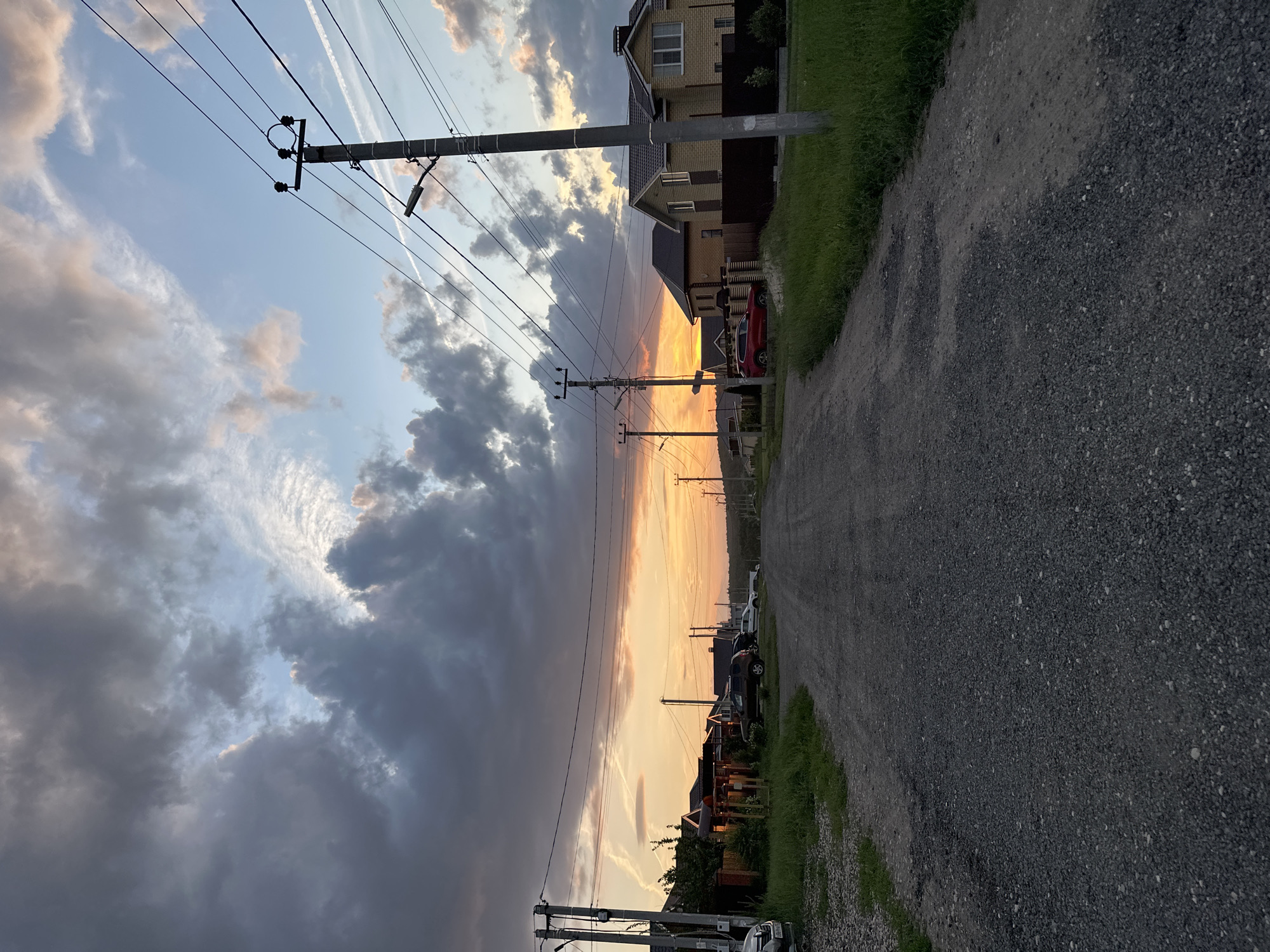 The image depicts a rural road in the city of Estachi, Russia, during a sunset. A gravel road stretches into the distance, flanked by grassy verges and houses on either side. Several cars are parked along the road and in driveways. Power lines and utility poles are visible throughout the scene, with a street light attached to one of the poles. The sky is filled with dramatic clouds, ranging from dark grays to lighter pinks and oranges, indicative of a sunset. Contrails from airplanes are faintly visible in the sky. The overall impression is of a quiet, residential area at the end of the day.