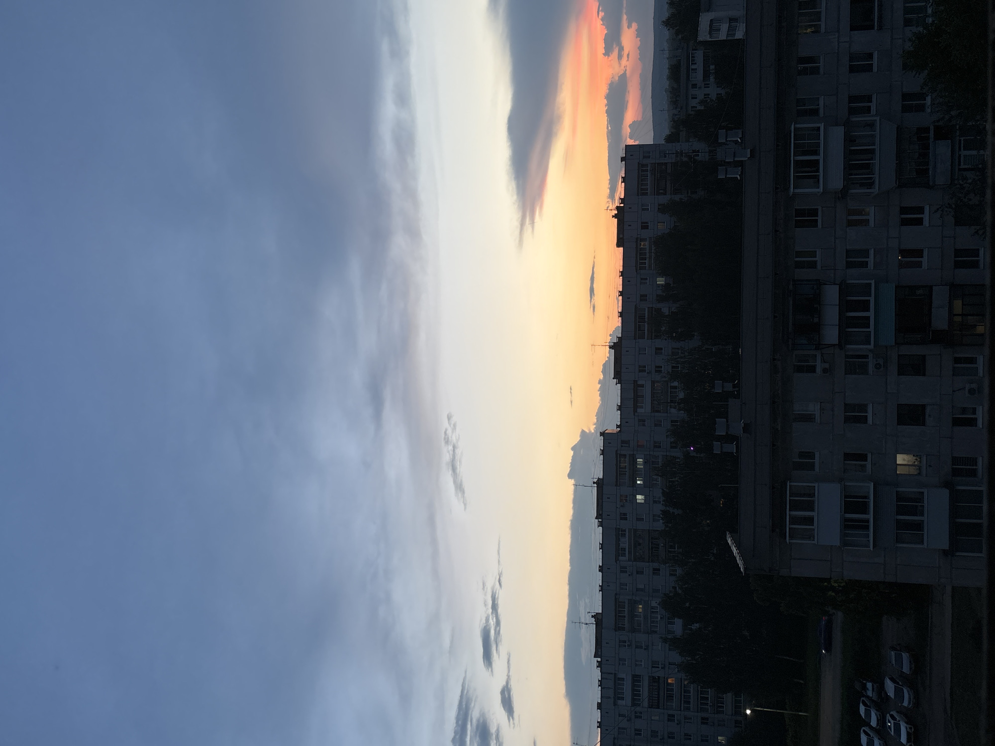 This is a vertical, high-angle photograph of an urban landscape at dusk in Chaikovsky, Russia. The sky is filled with a gradient of colors, transitioning from a deep blue at the top to shades of gray and white, with a vibrant band of orange and pink near the horizon. Clouds are scattered across the sky, some thin and wispy, others denser and more defined.
In the foreground, two multi-story apartment buildings dominate the scene. The closer building, on the bottom, is dark and appears to be made of concrete, with numerous windows. Some windows are illuminated, suggesting interior lights are on. The building behind it is larger and taller, also appearing to be constructed of concrete. It has a more complex facade with visible balconies and window arrangements. Trees line the base of this taller building, casting dark shadows.
To the left of the apartment buildings, several cars are parked in what appears to be a parking lot. The cars are a mix of white, silver, and dark colors. A street light is visible on the left edge of the frame, casting a subtle illumination. In the far distance, a faint outline of hills or a low mountain range can be seen against the colorful sky. The overall impression is one of a quiet evening in a residential area.