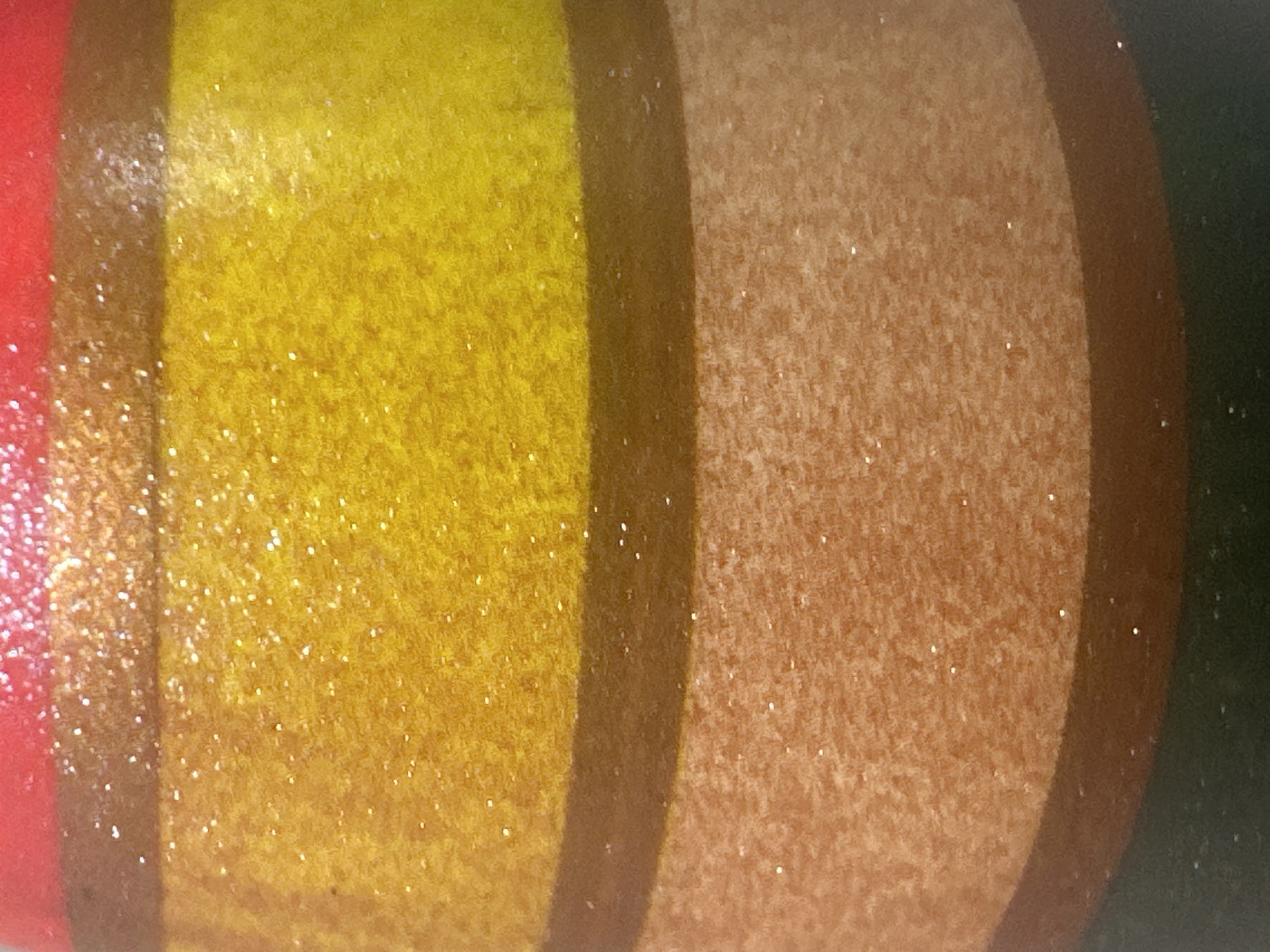 This is a close-up, detailed, abstract shot of a surface with horizontal stripes of color. The top stripe is red, followed by a thin brown stripe, then a wide yellow stripe with glitter, another thin brown stripe, and a wide tan stripe with glitter. The bottom of the image shows a dark green stripe. The image does not depict any elements that would allow for the identification of a specific location in Letterkenny, Ireland.