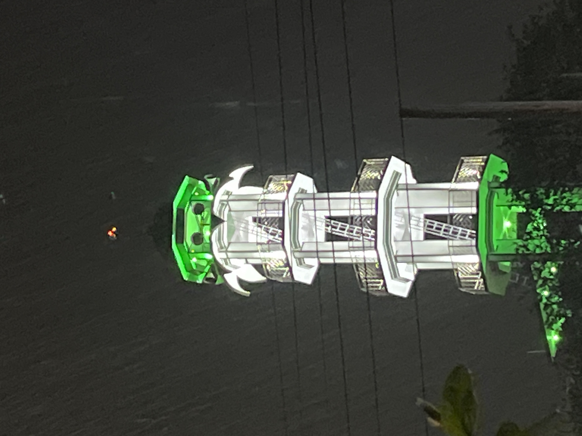 The image captures a brightly illuminated ferris wheel at night, likely located in Dhaka, Bangladesh. The ferris wheel is decorated with white and green lights and has a complex, multi-tiered structure. Power lines are visible in the foreground, suggesting an urban setting. The surrounding environment is dark, possibly indicating a park or other outdoor venue at night. In the distance, there are a couple of orange glowing orbs, likely other sources of light. There are also trees visible.